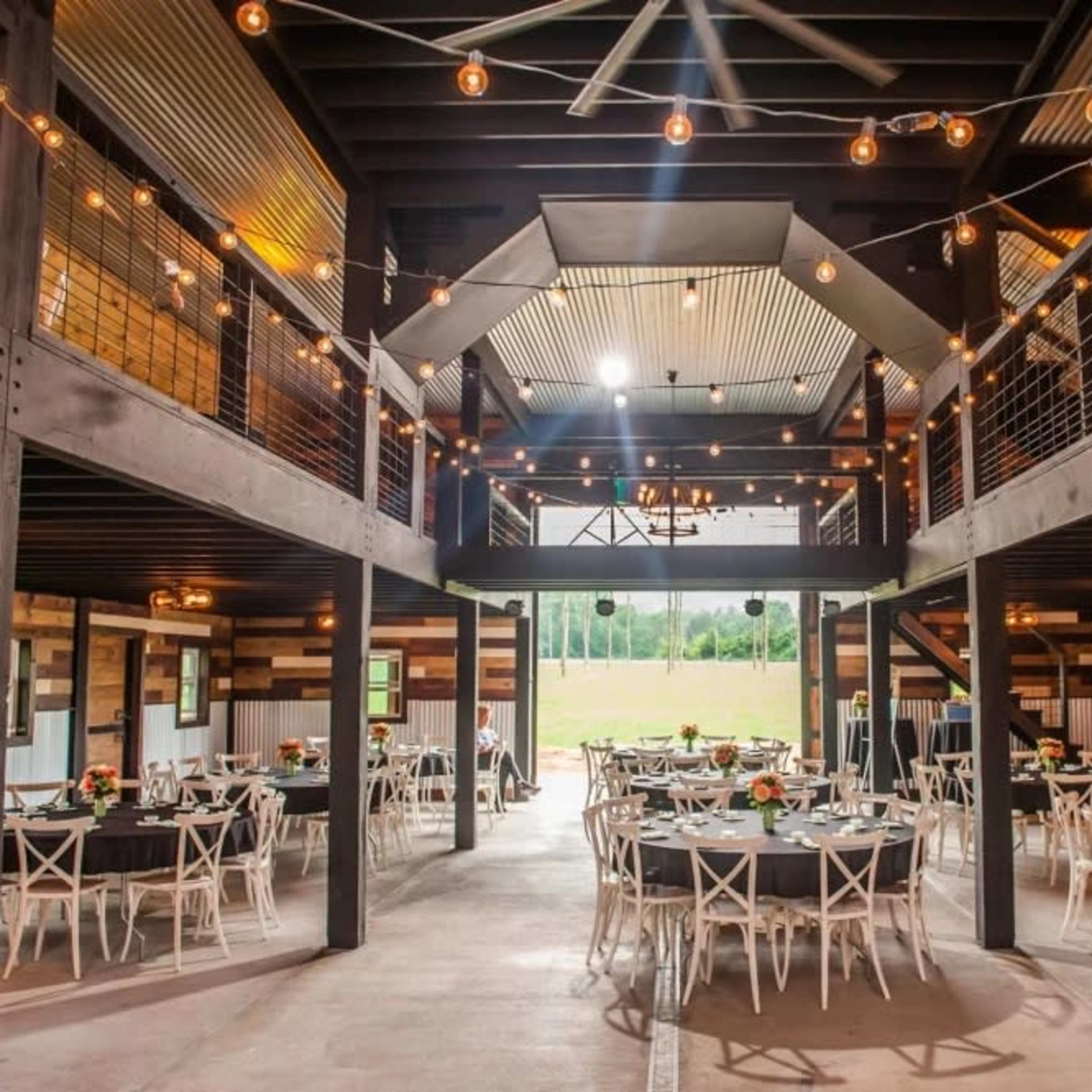 A spacious event venue with round tables set for dining, string lights overhead, and a two-level design featuring an open layout.