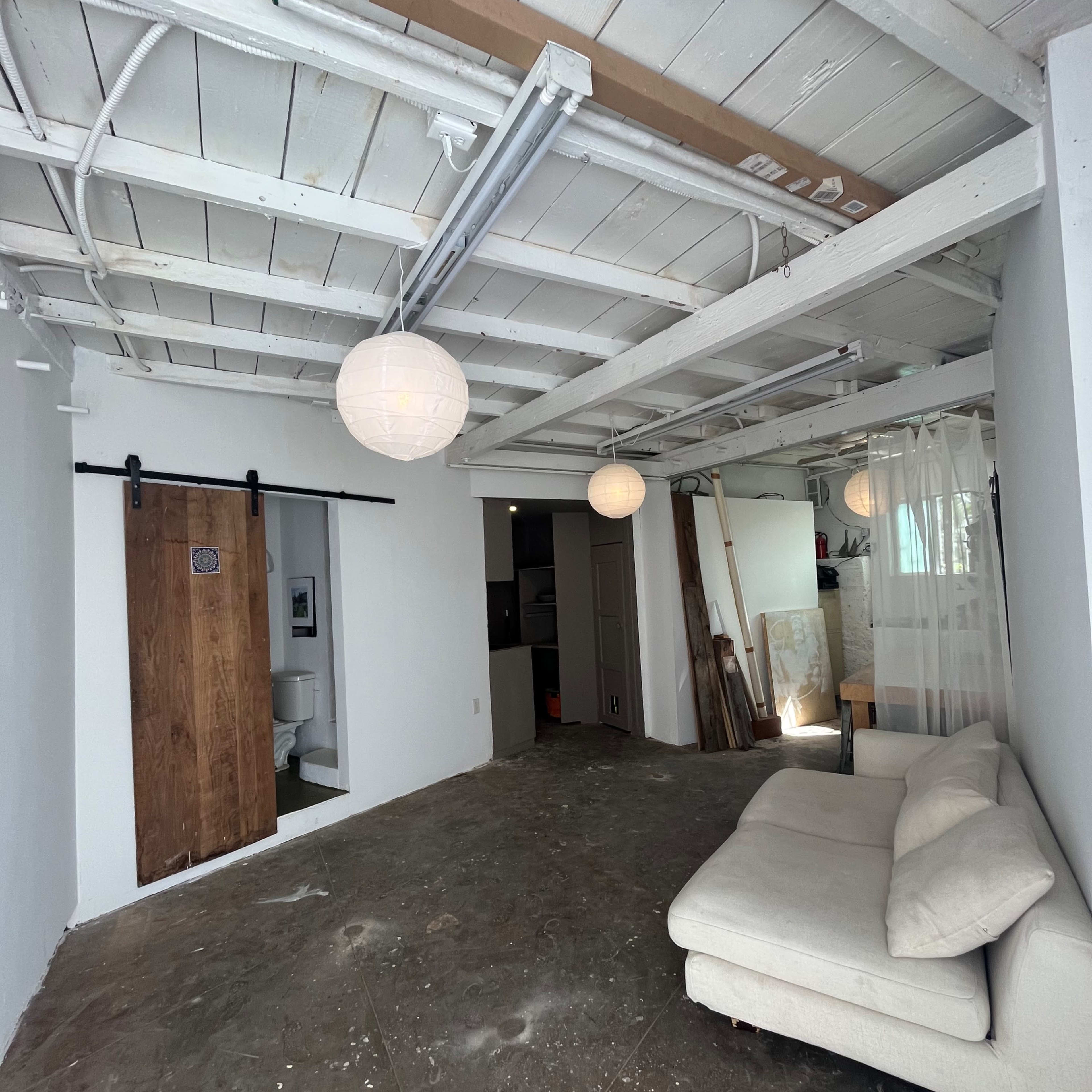 A minimalist interior space features a white sofa, exposed beams, hanging light fixtures, and a wooden sliding door leading to a bathroom.
