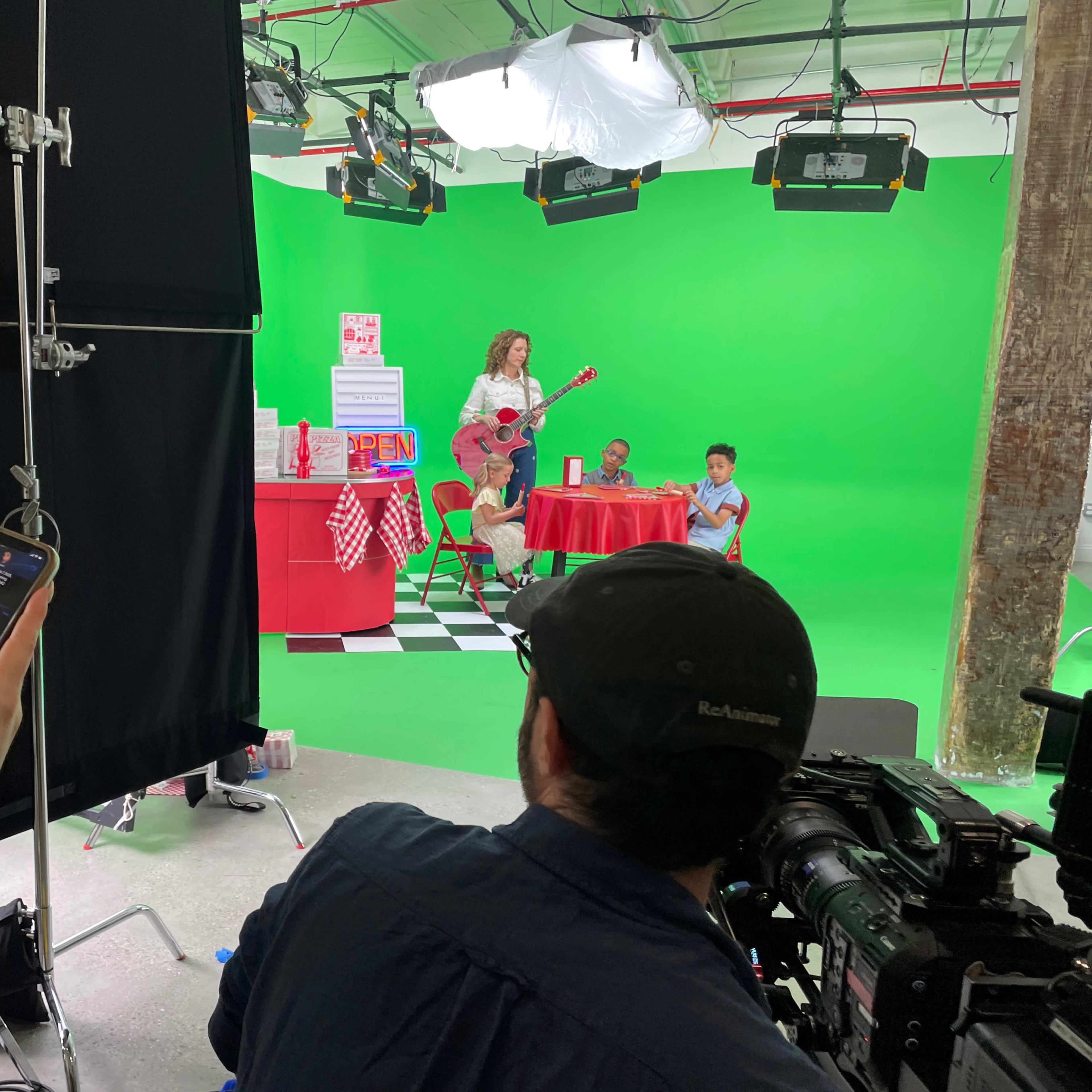 A crew member holds a camera while another uses a phone to monitor a scene being filmed in front of a green screen, featuring a woman playing guitar and two children seated at a table adorned with promotional items.