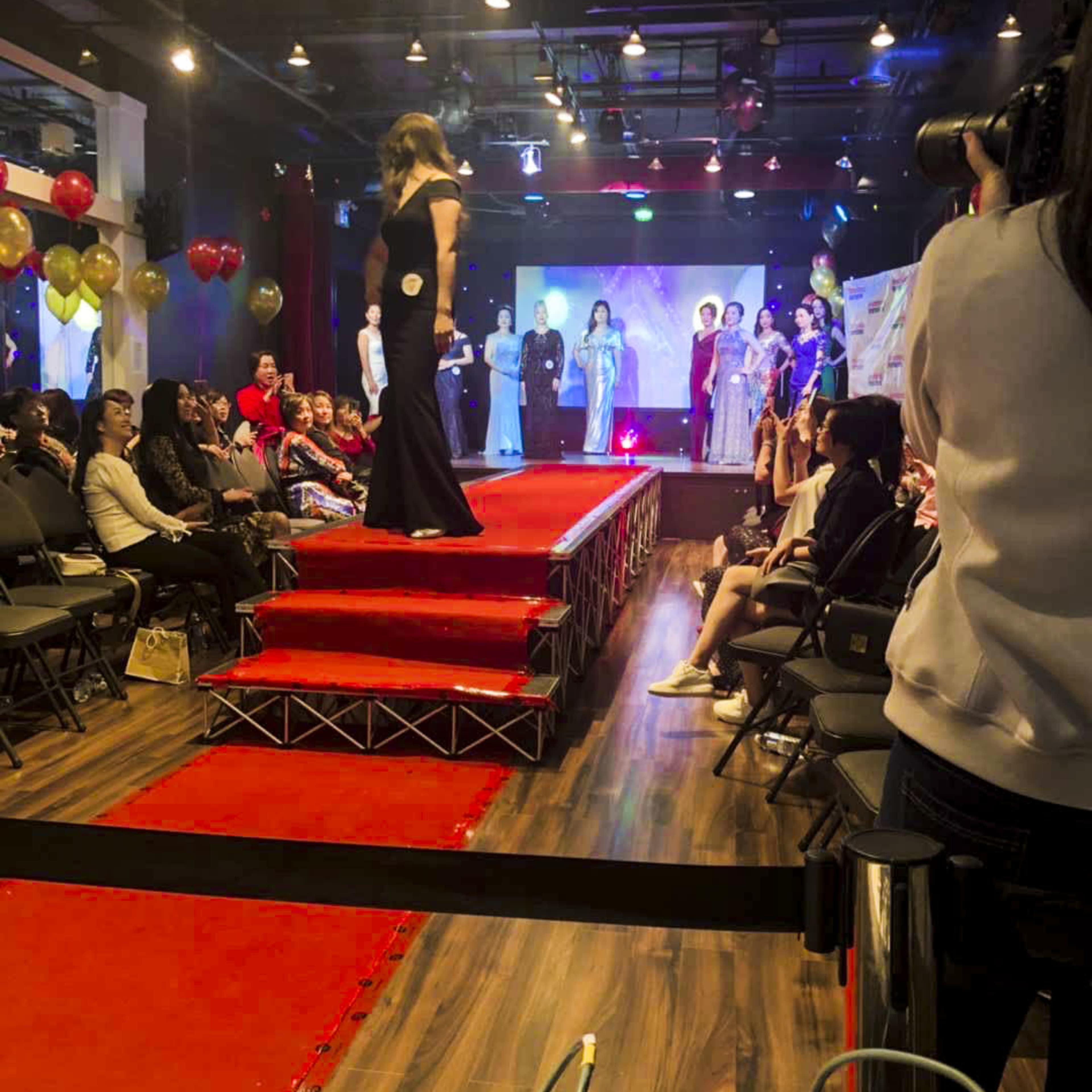A fashion runway features models showcasing garments on a red carpet while an audience watches from seated rows.