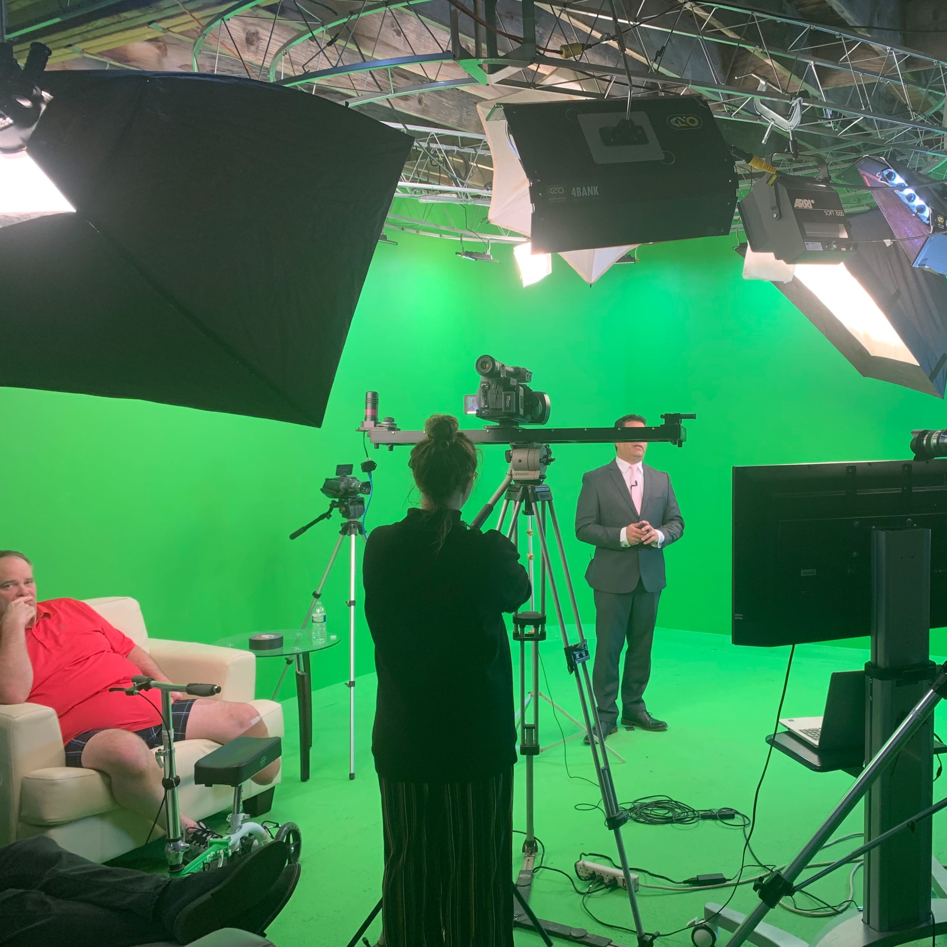 A man in a suit stands in front of a green screen, surrounded by cameras and lights, while a crew prepares to film.