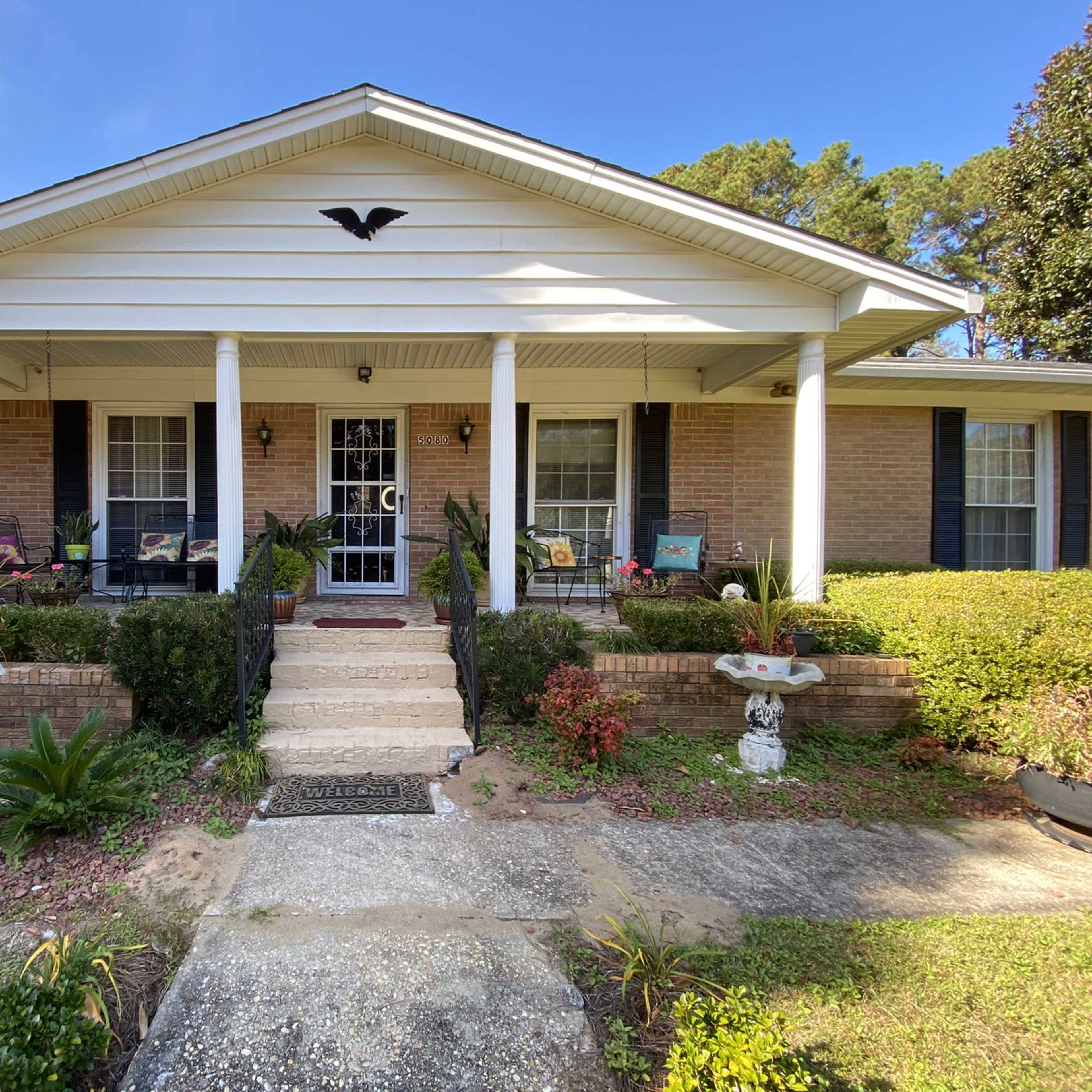 A single-story brick house with a front porch, white columns, and well-maintained landscaping.