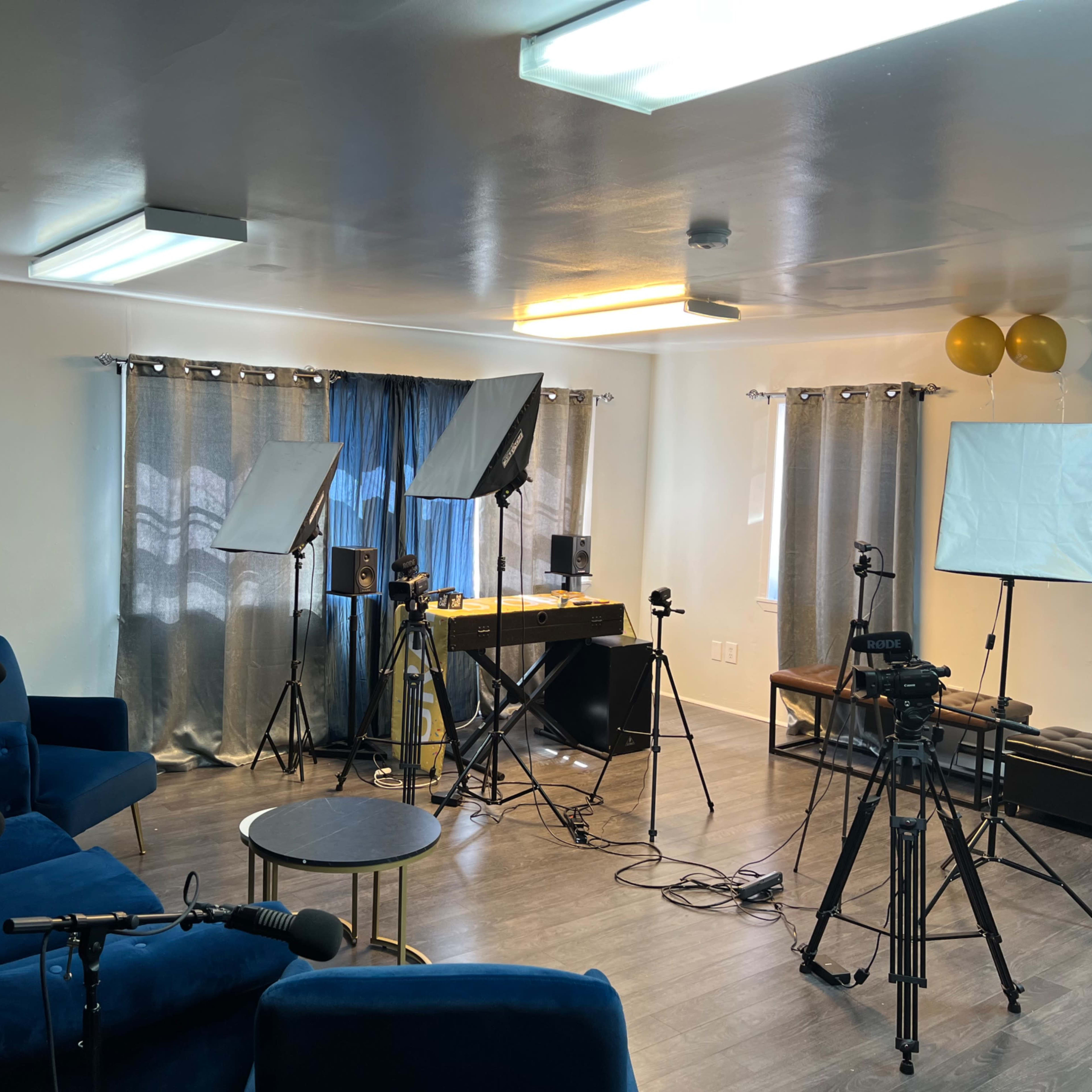 The room is set up for filming, with blue furniture, lighting equipment, and multiple cameras positioned around a table.