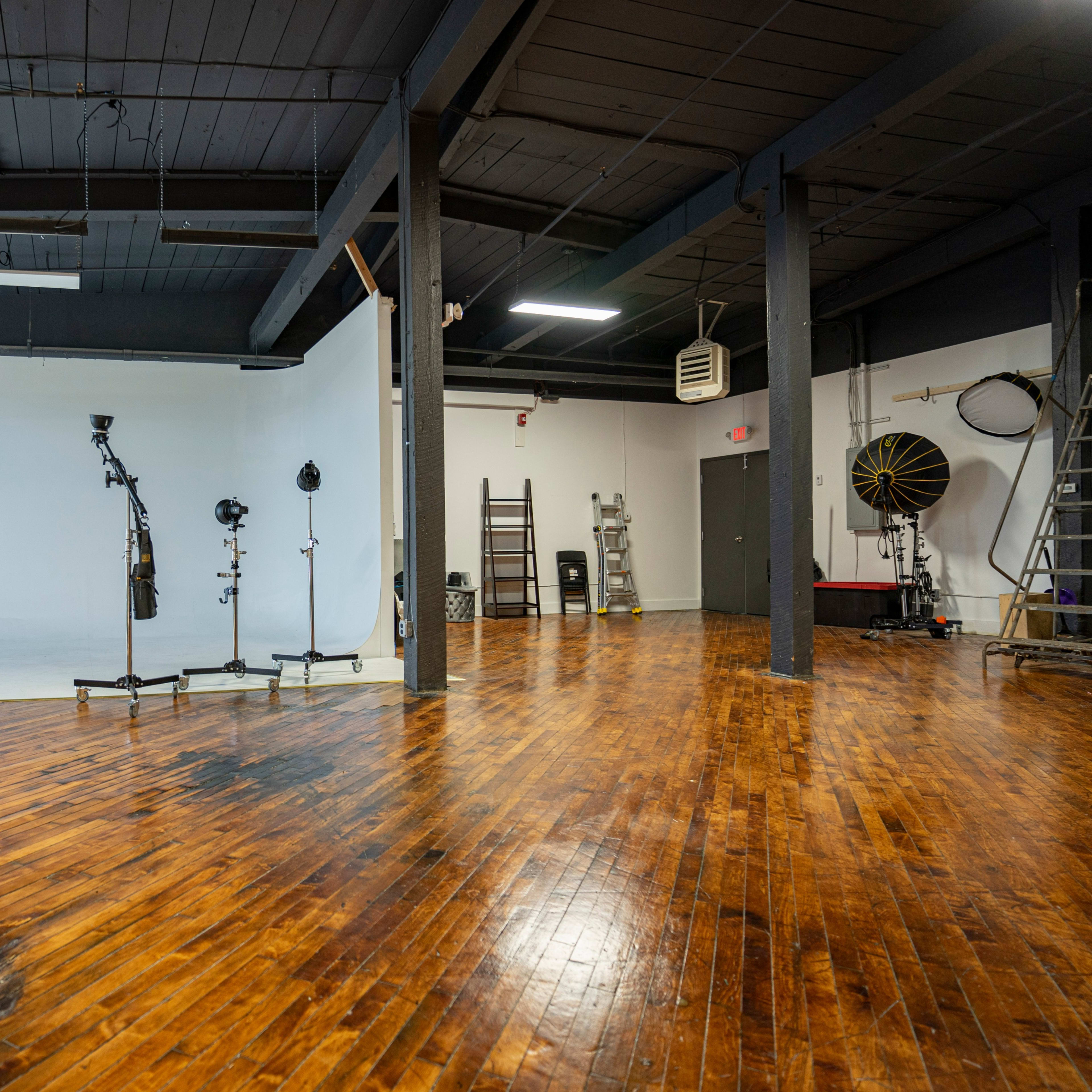 A spacious photography studio with a white backdrop, wooden flooring, and various lighting equipment arranged in the area.