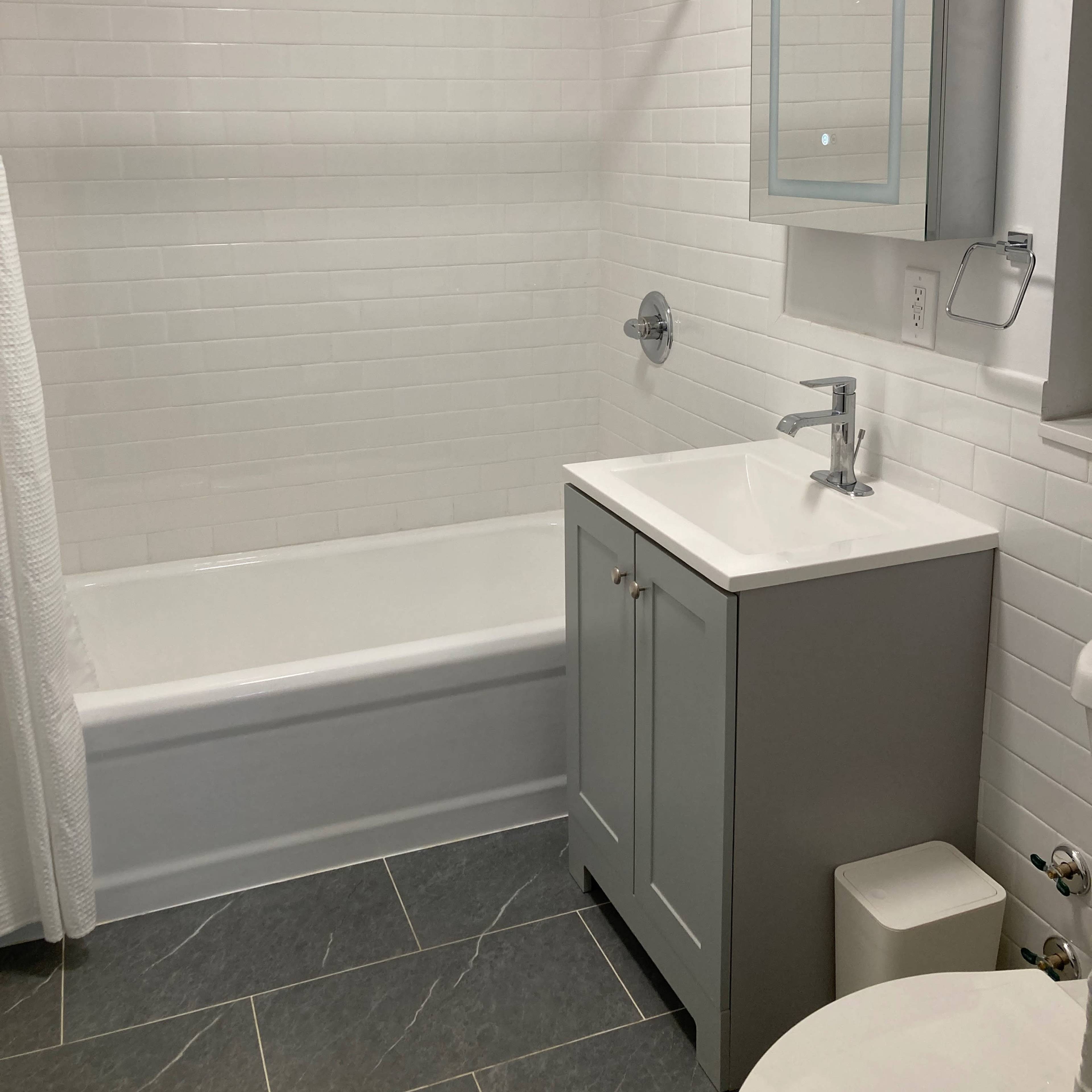 A modern bathroom with a bathtub, a gray vanity with a sink, a wall-mounted mirror, and white tiled walls.