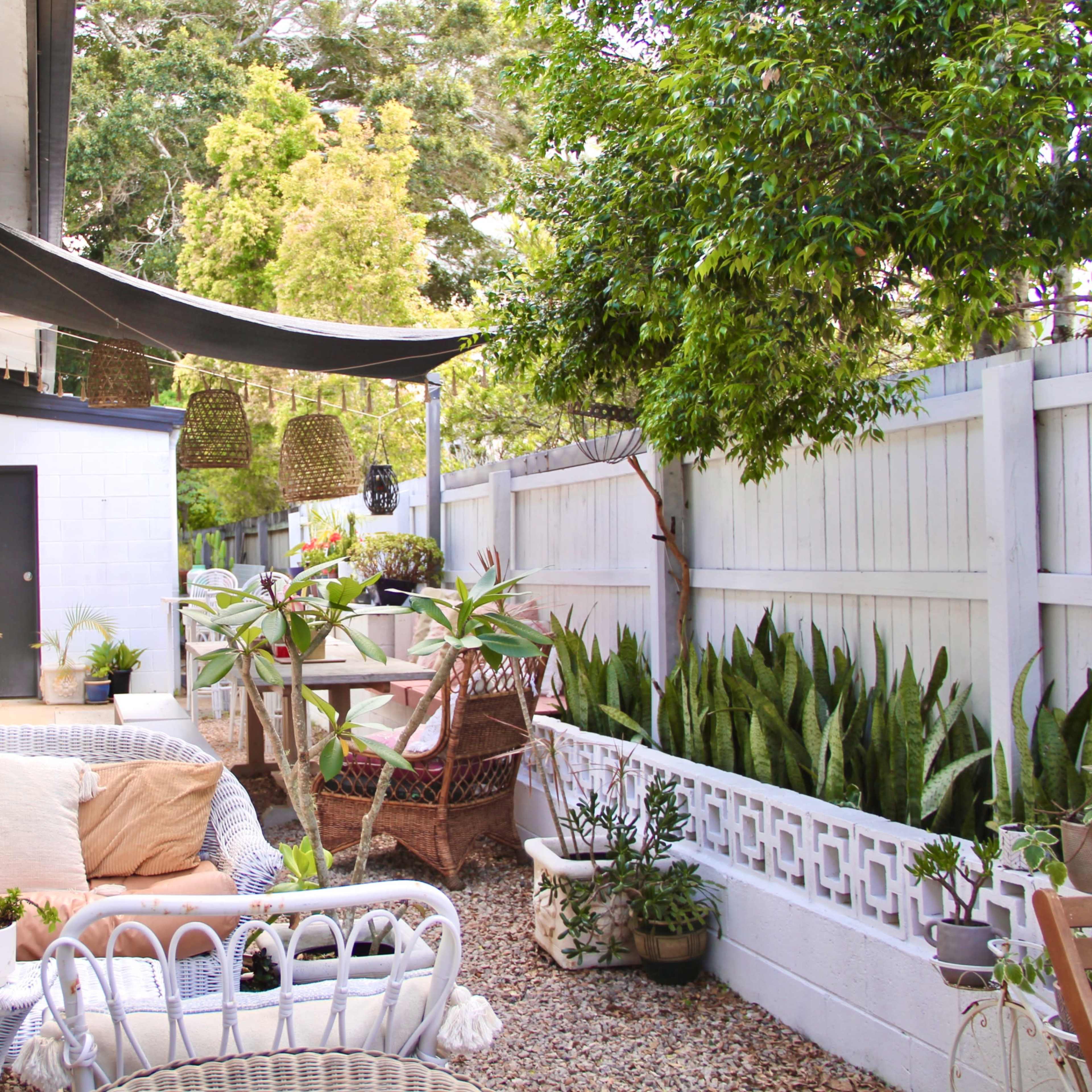 A patio area with wicker furniture, potted plants, and a shaded dining table surrounded by greenery and a wooden fence.