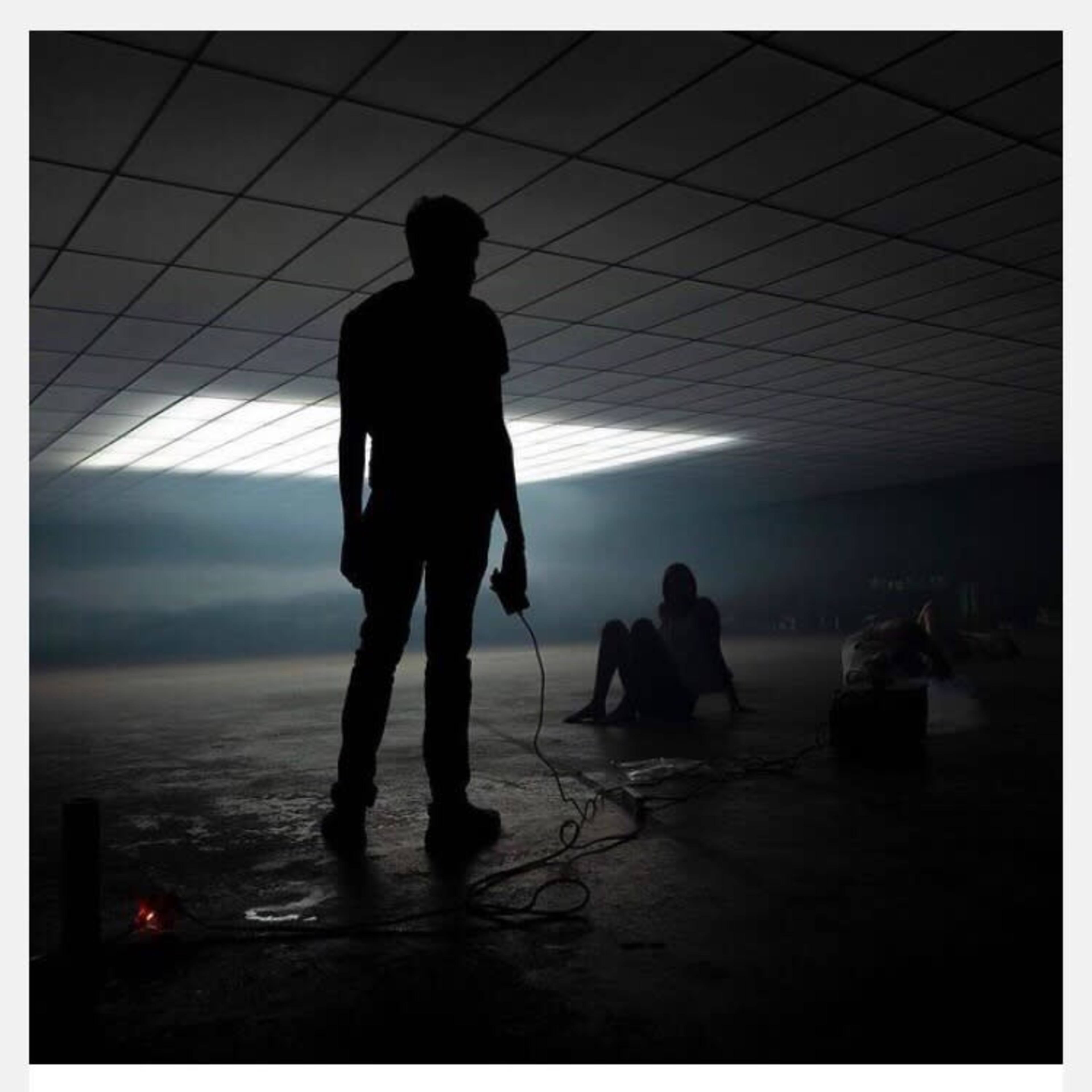 A silhouette of a person stands in a dimly lit room with another figure seated in the background, surrounded by beams of light and smoke.