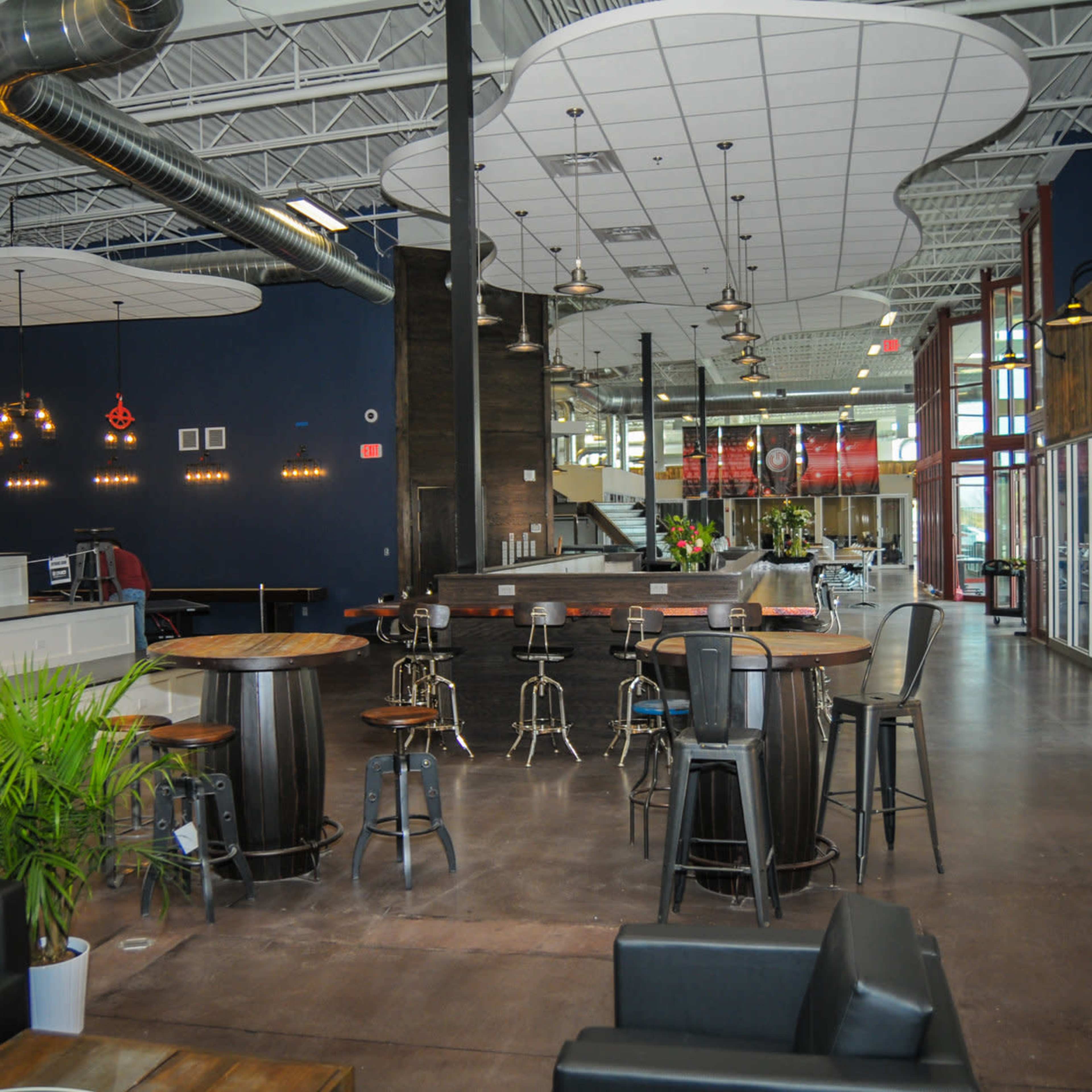 An expansive, modern interior of a café with round tables, bar stools, and a service counter, featuring high ceilings and large windows.