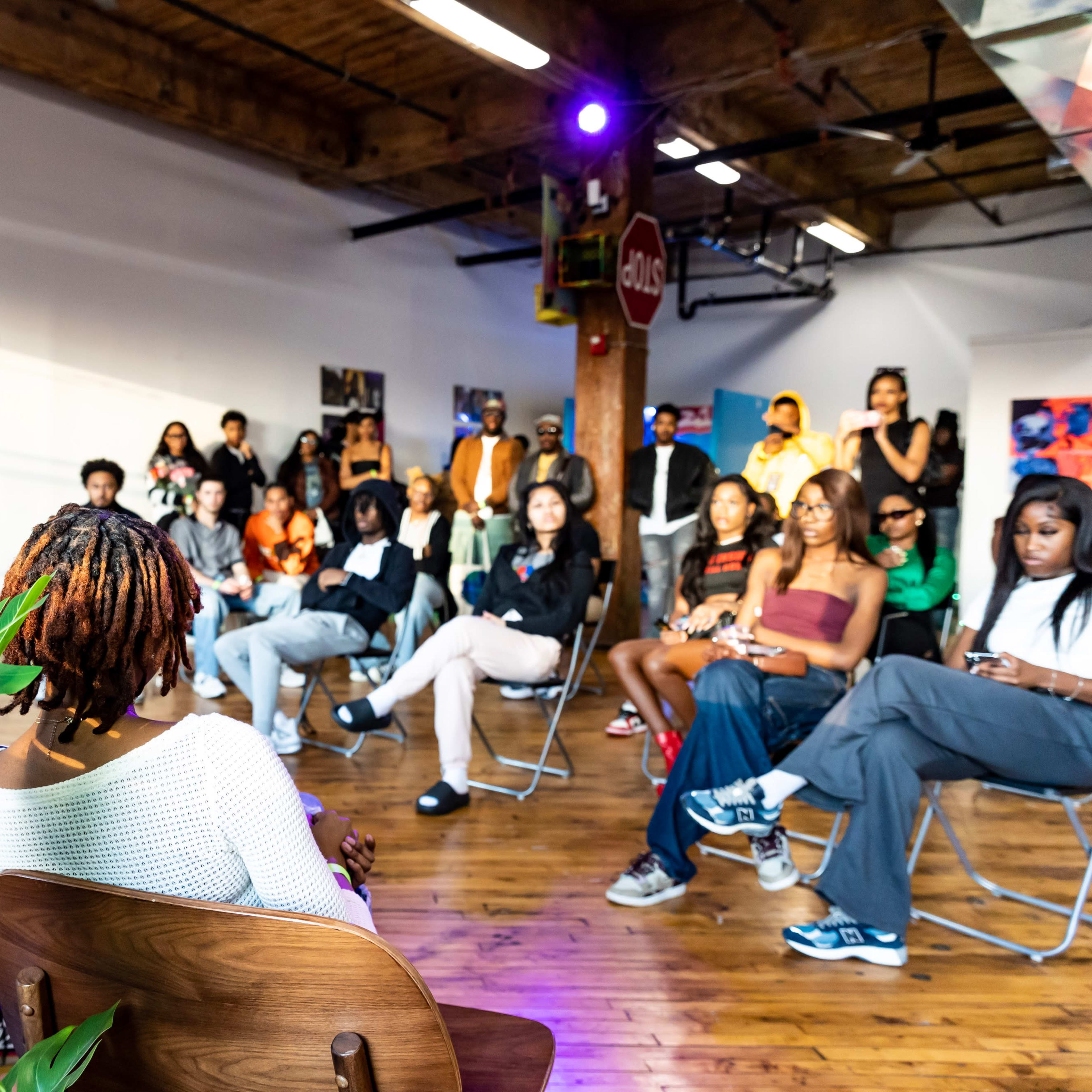 A diverse group of people sits in a gallery space, attentively listening to speakers at a community event.