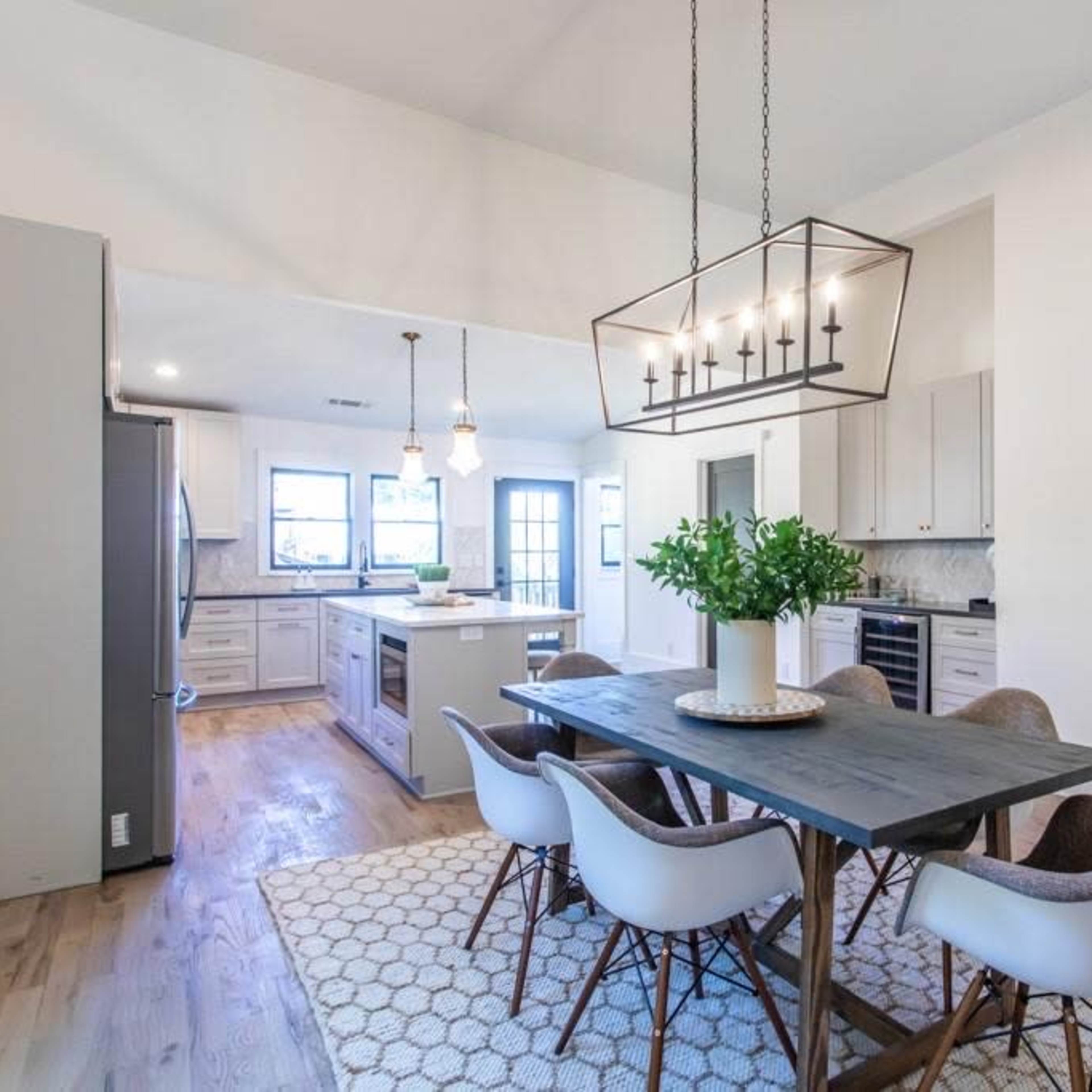 A modern kitchen and dining area features a wooden table with chairs, overhead lighting, and a large island with appliances.