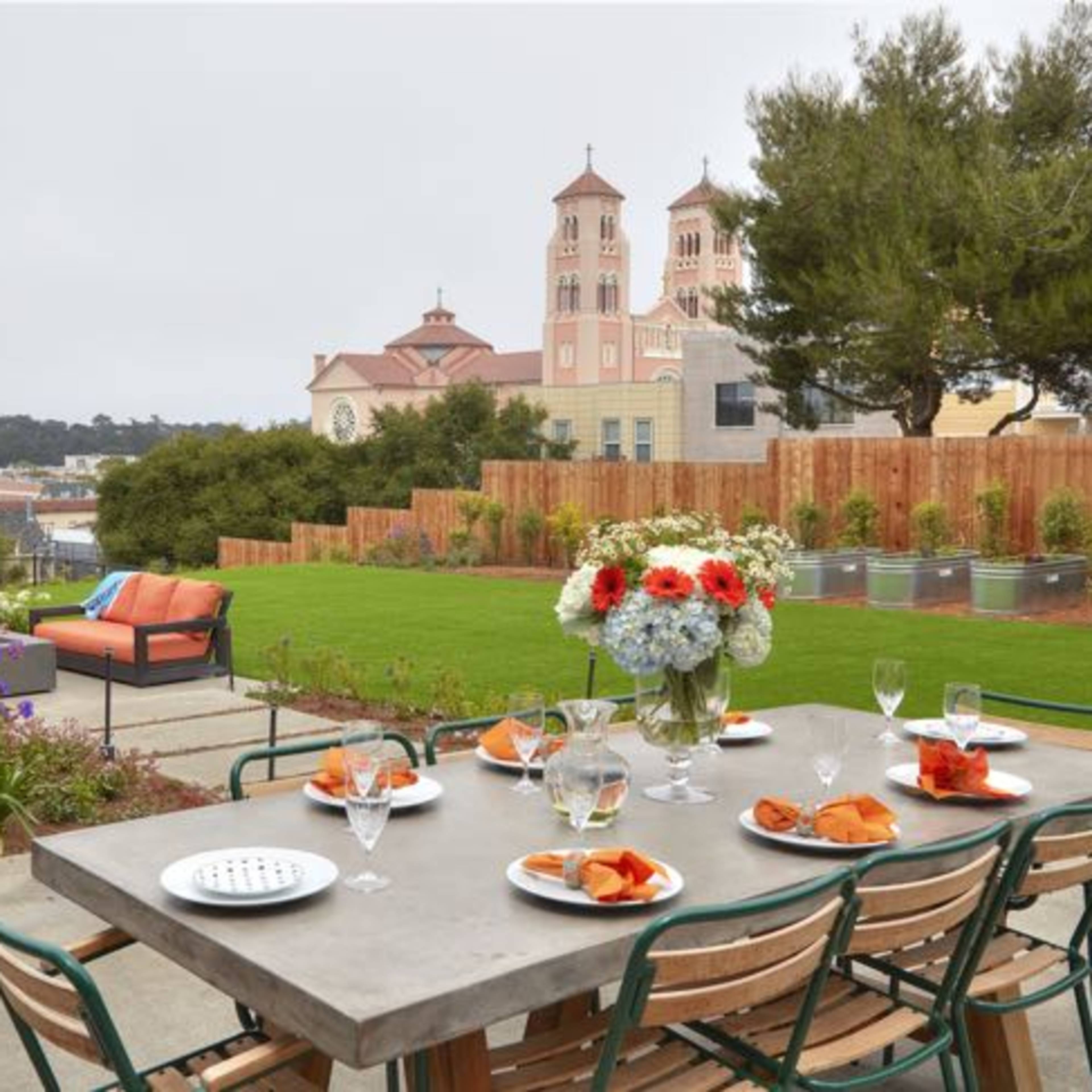 A dining table is set in a landscaped backyard with flowers and a view of a building with two towers in the background.