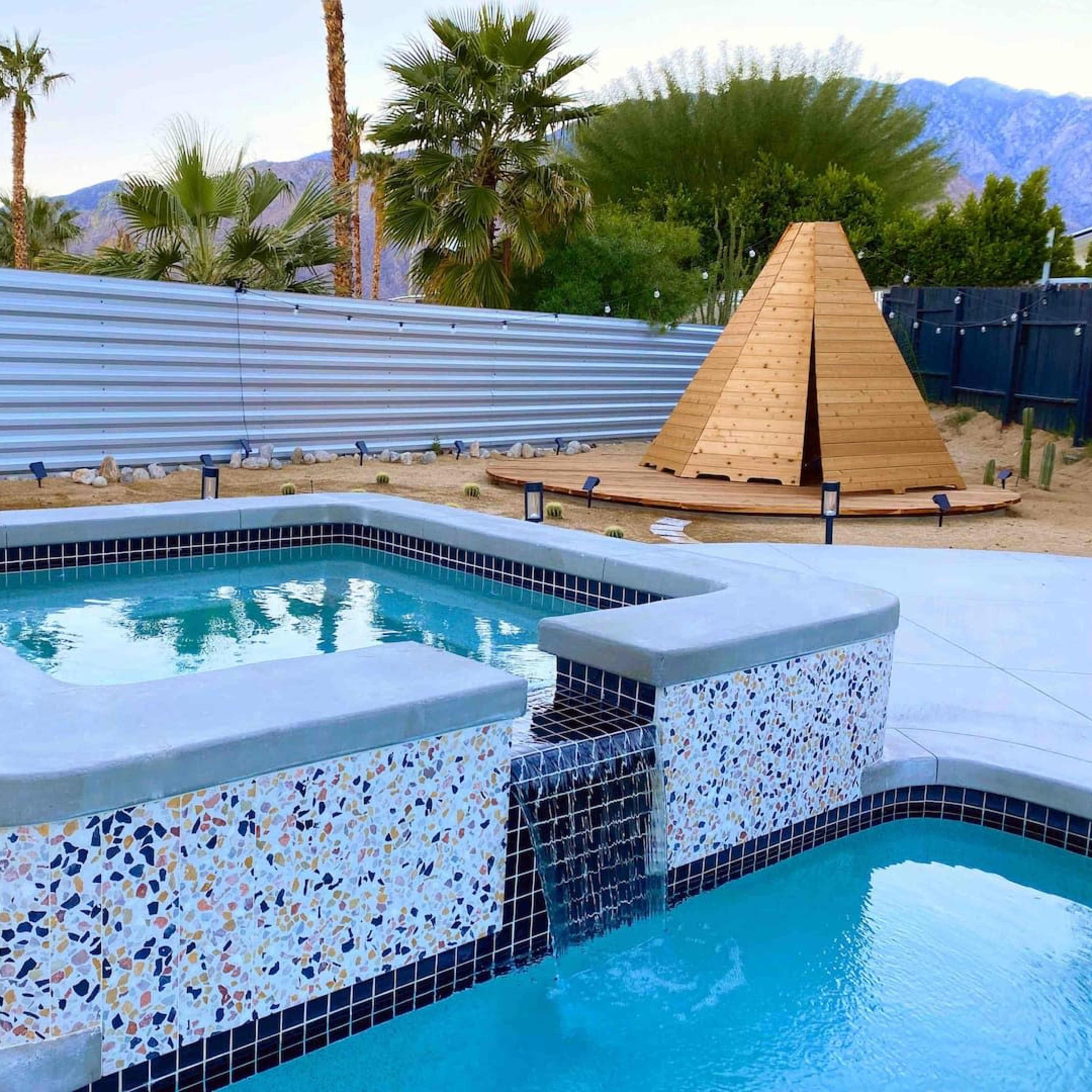 A backyard with a spa and a rectangular pool, surrounded by a fenced area and featuring a wooden pyramid structure in the background.