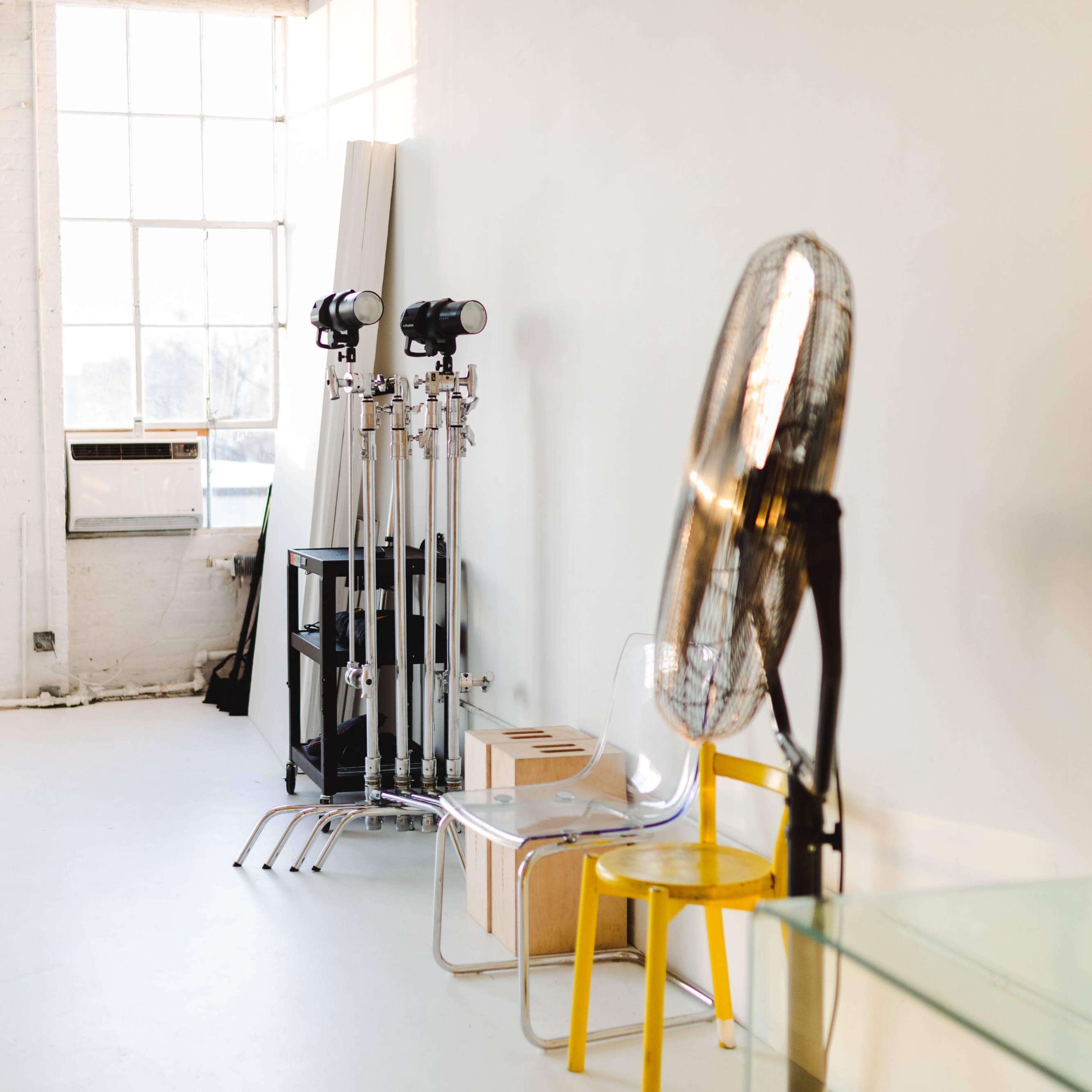 A bright, minimalist studio space with equipment stands, a fan, and a chair against a wall of large windows.