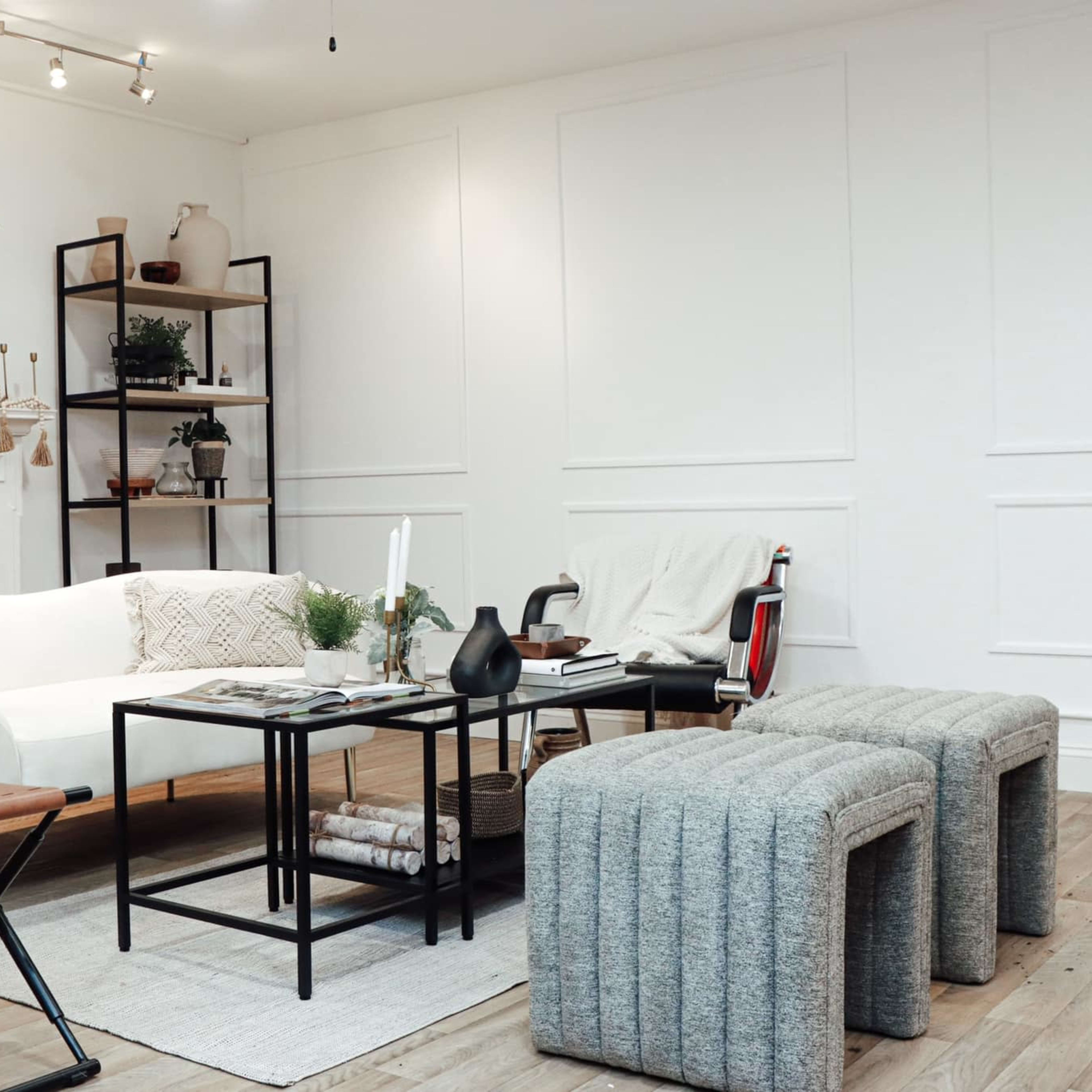 A bright living room featuring a white sofa, a black coffee table, and two gray upholstered stools, with shelves and plants in the background.