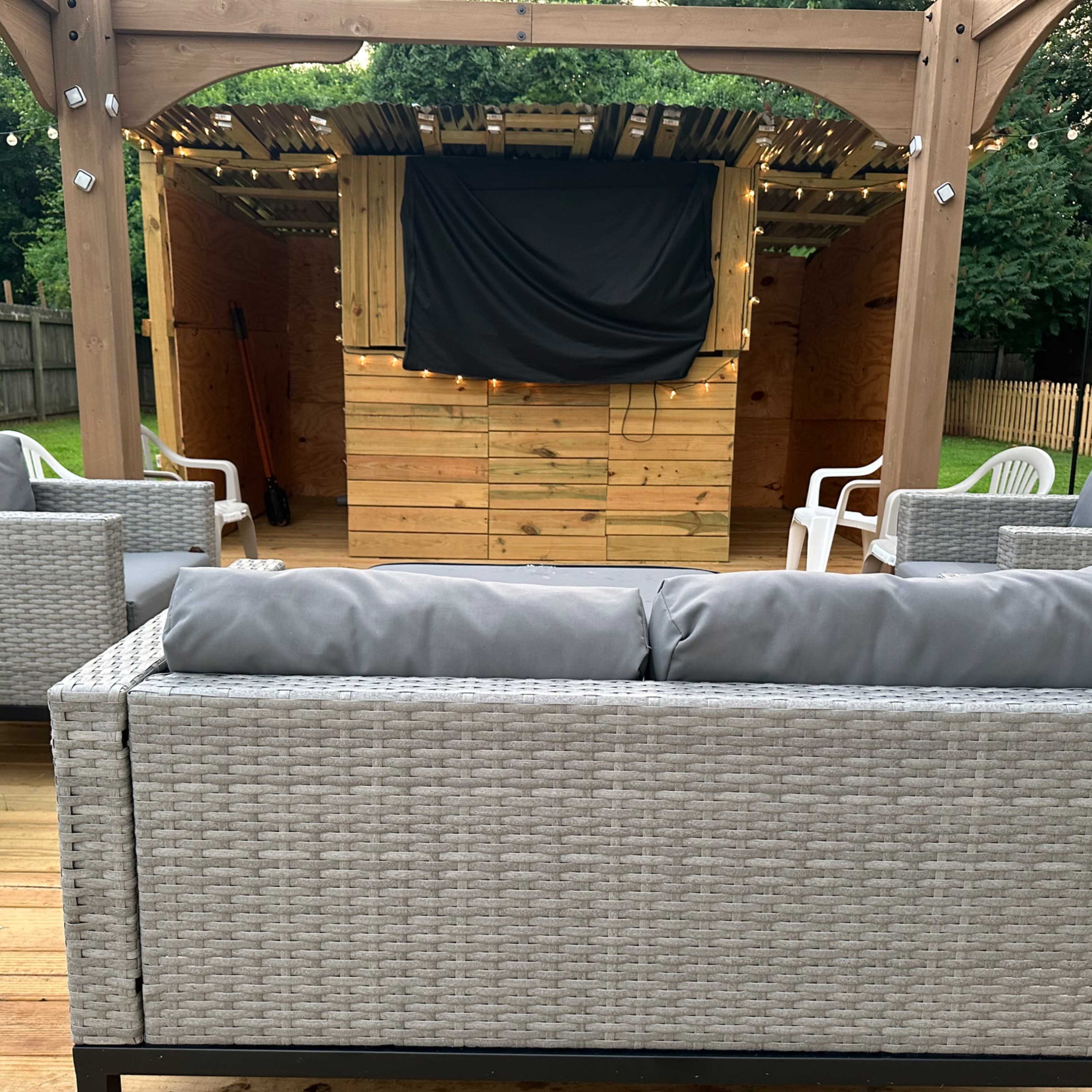 A patio area with several outdoor seating arrangements facing a wooden stage covered by a black cloth, illuminated by string lights overhead.