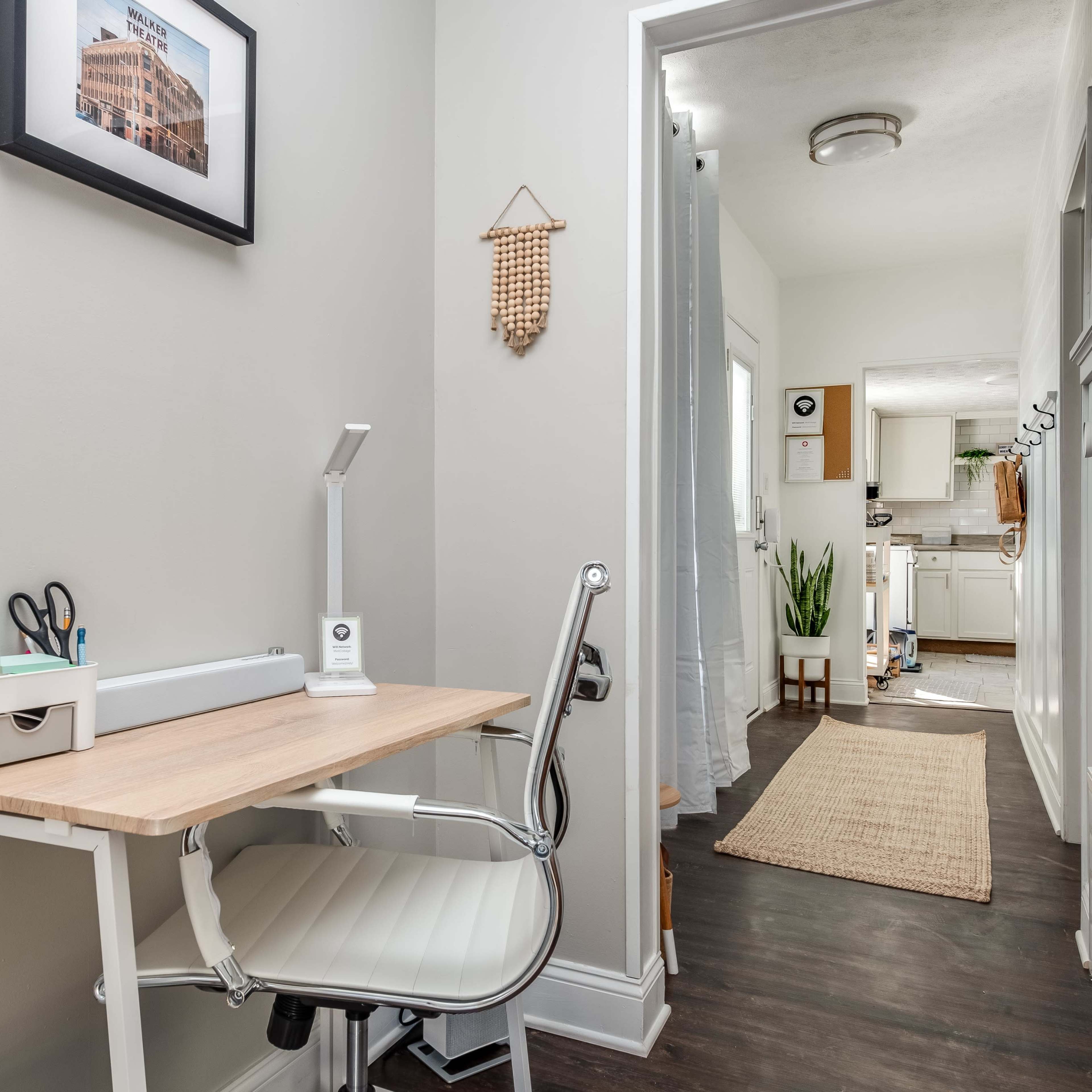 A bright hallway featuring a small workstation with a white chair and wooden desk, leading to a well-lit kitchen area.