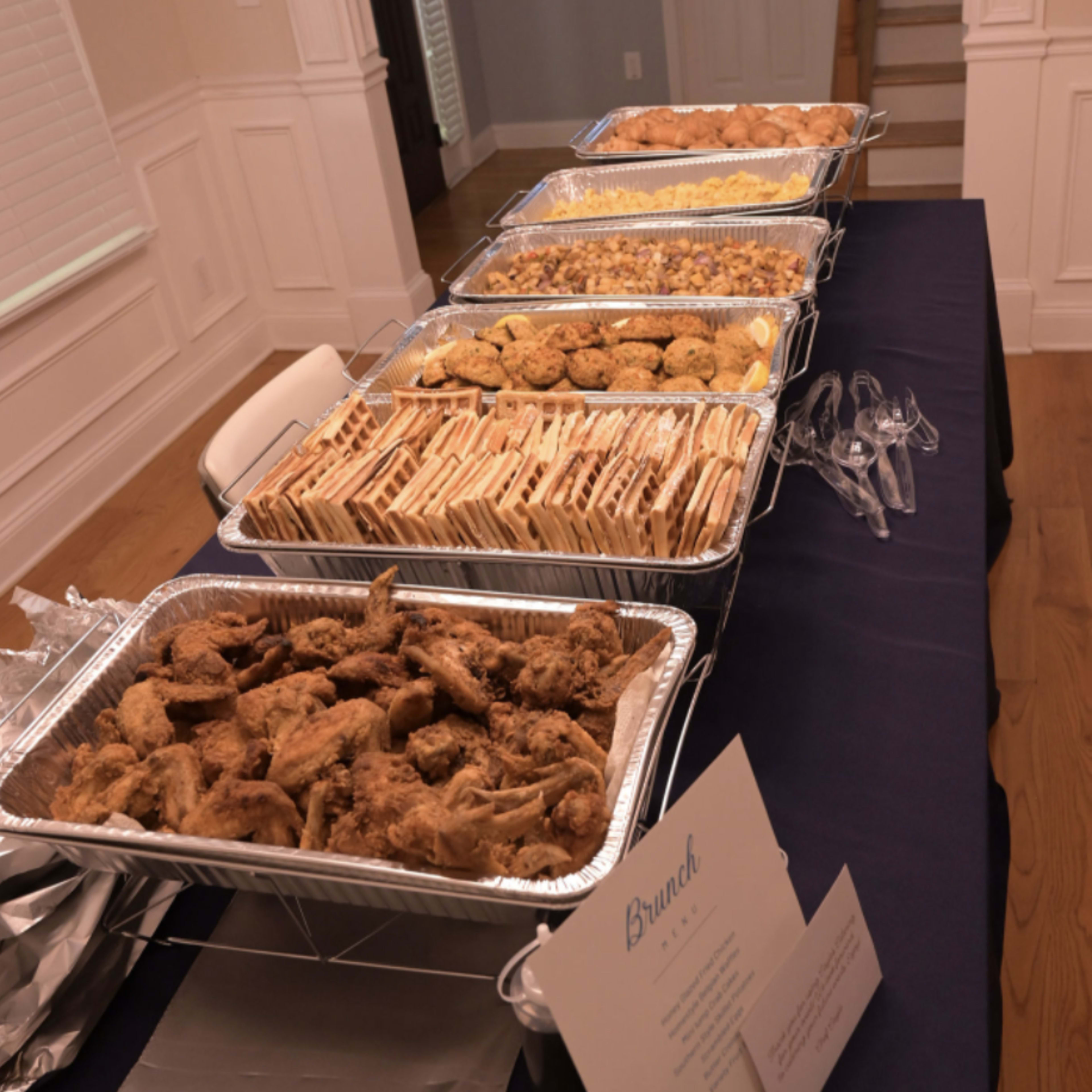 A table is set up with several trays of food for a brunch, featuring items such as fried chicken, pastries, and a variety of baked goods, accompanied by a decorative sign.
