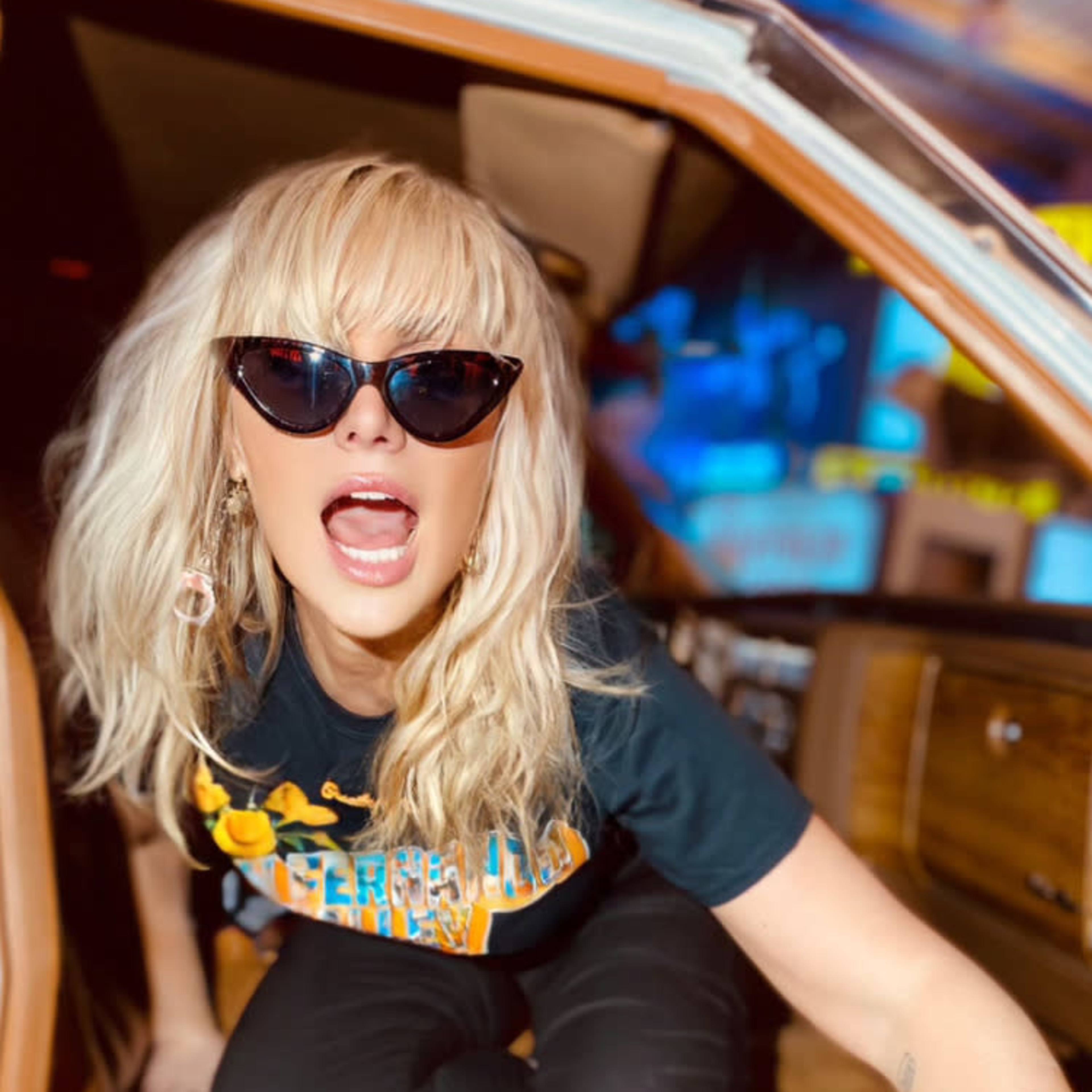 A woman with blonde hair and sunglasses is sitting in a vintage car, leaning out of the driver's side.