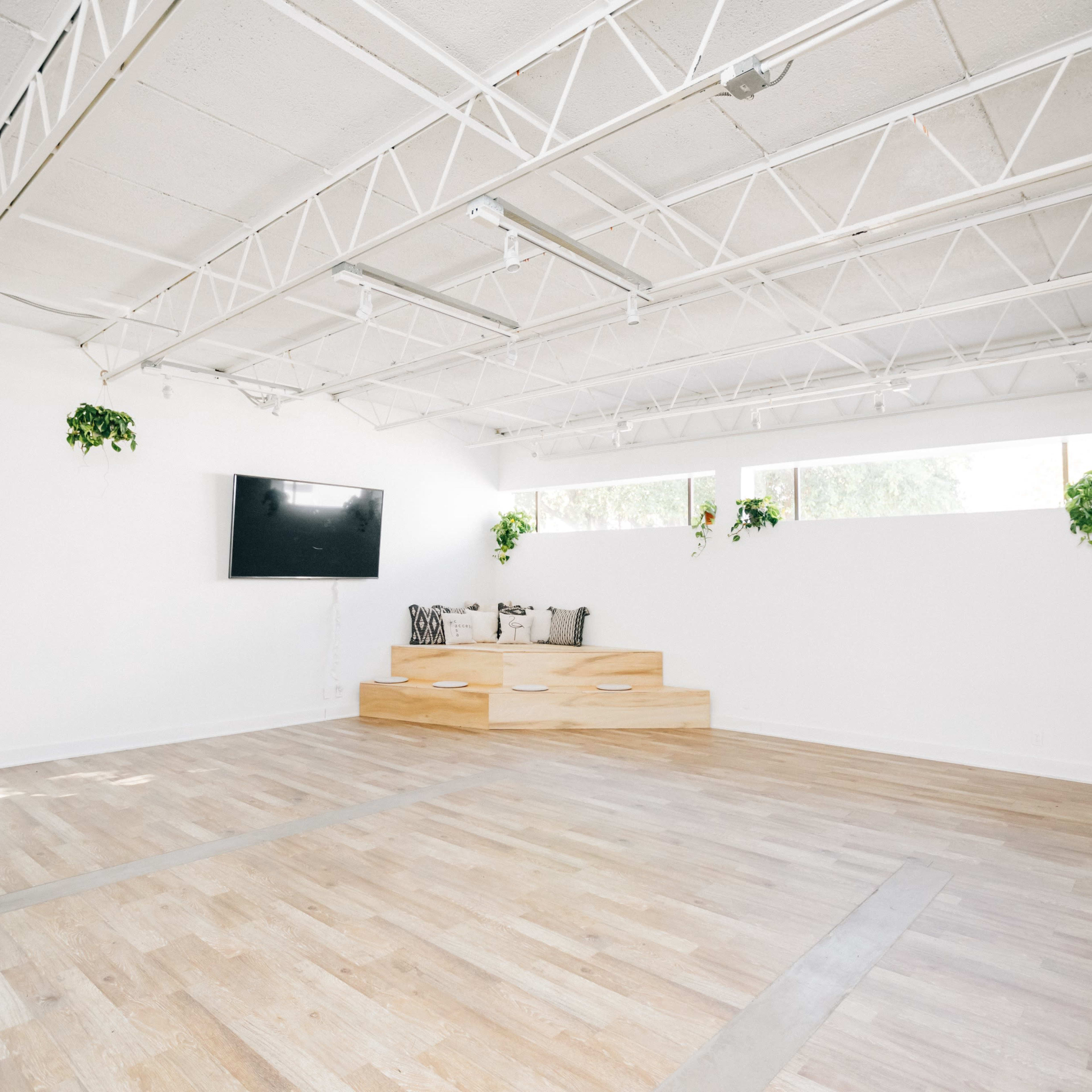 A spacious, empty room with wooden flooring, a large television mounted on the wall, and a raised wooden platform with seating by the windows.
