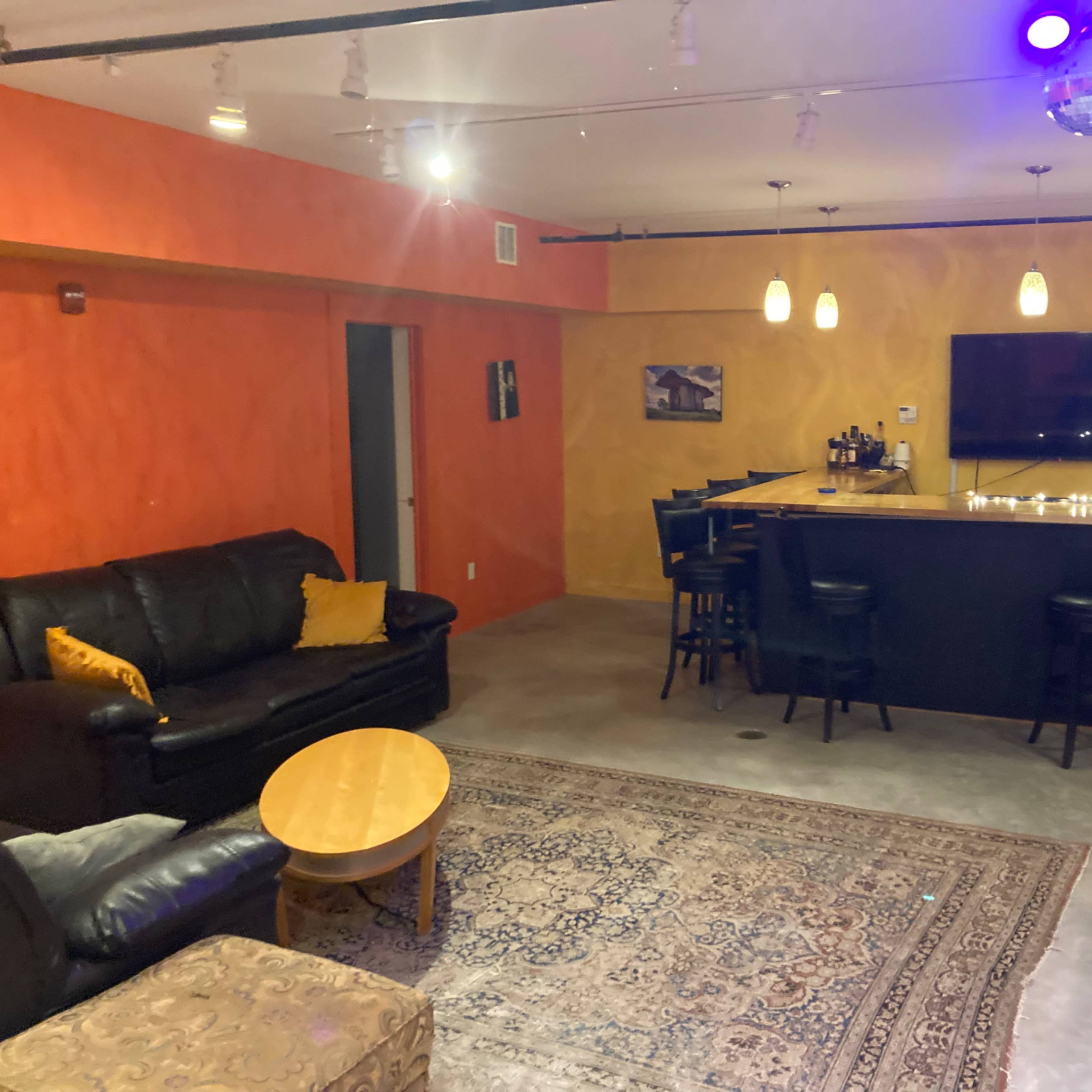 A colorful lounge area featuring a black leather couch, a round wooden coffee table, a bar with high stools, and a pool table, all illuminated by ambient lighting.