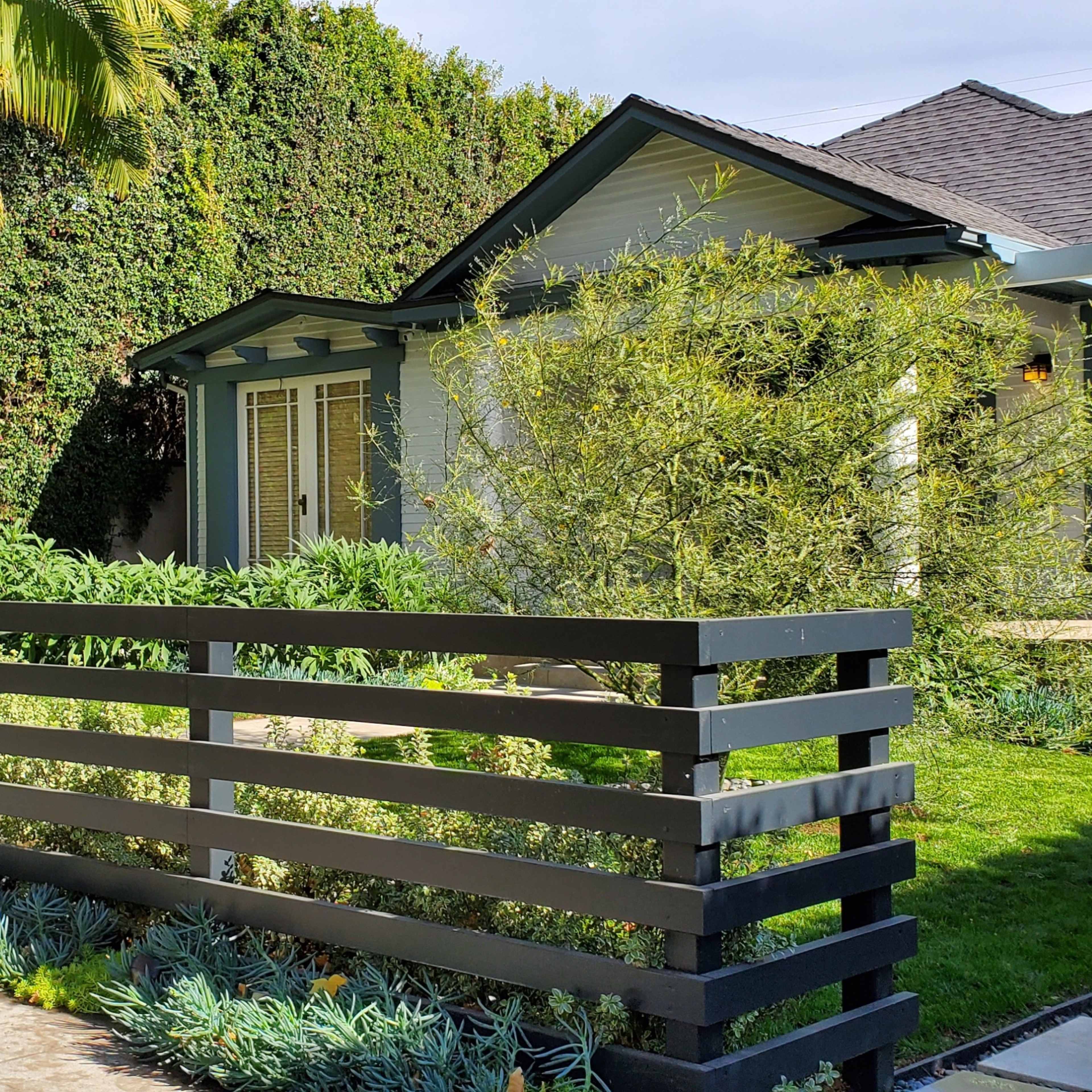 A landscaped front yard with a modern fence and a light-colored house surrounded by trees and shrubs.