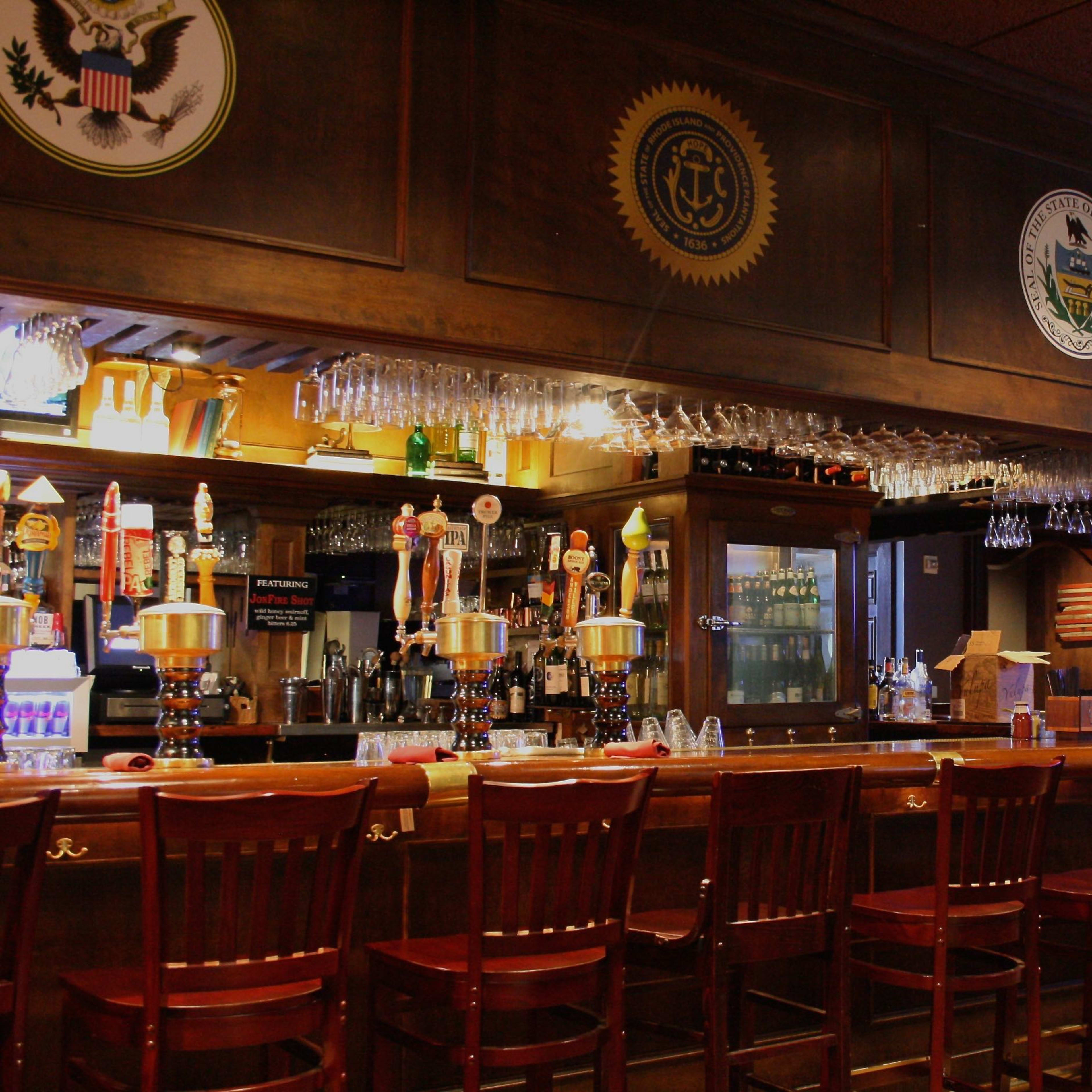 A wooden bar with several beer taps and a row of high-backed chairs, illuminated by soft lighting and decorated with emblems and flags on the walls.