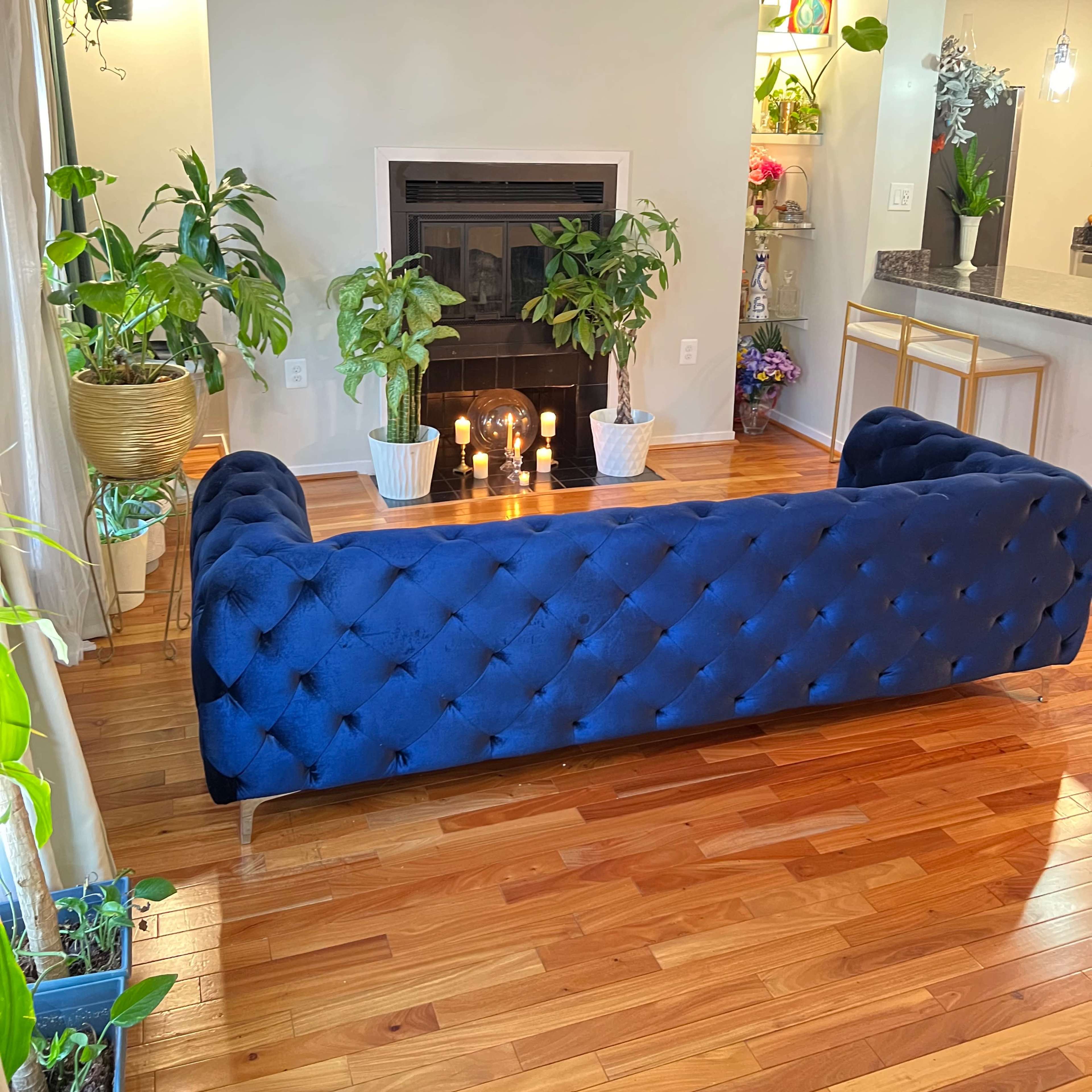 A blue tufted sofa faces a fireplace surrounded by potted plants and candles on a wooden floor.