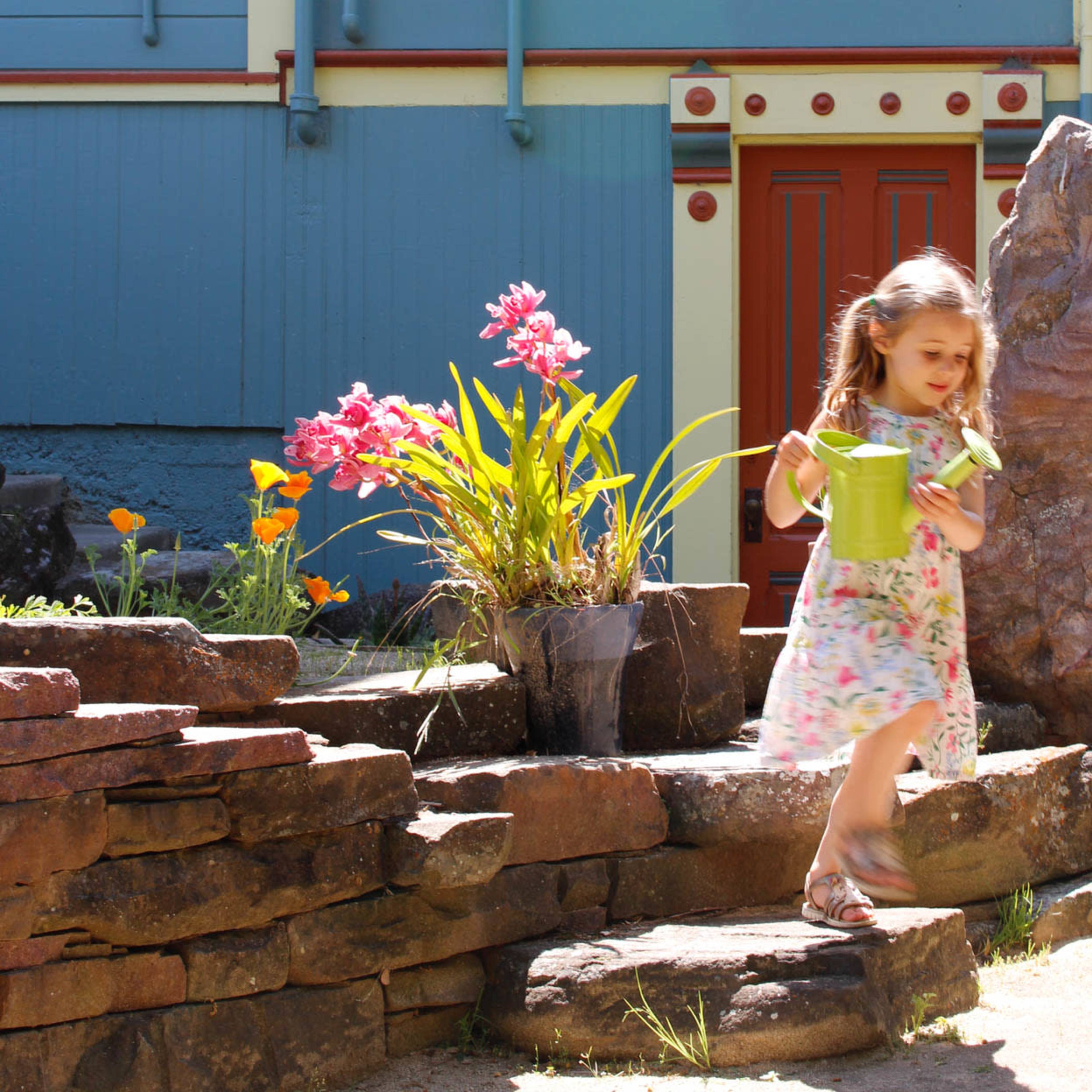 A young girl carries a green bucket while walking past a flower garden and a wooden map of California mounted on a colorful wall.