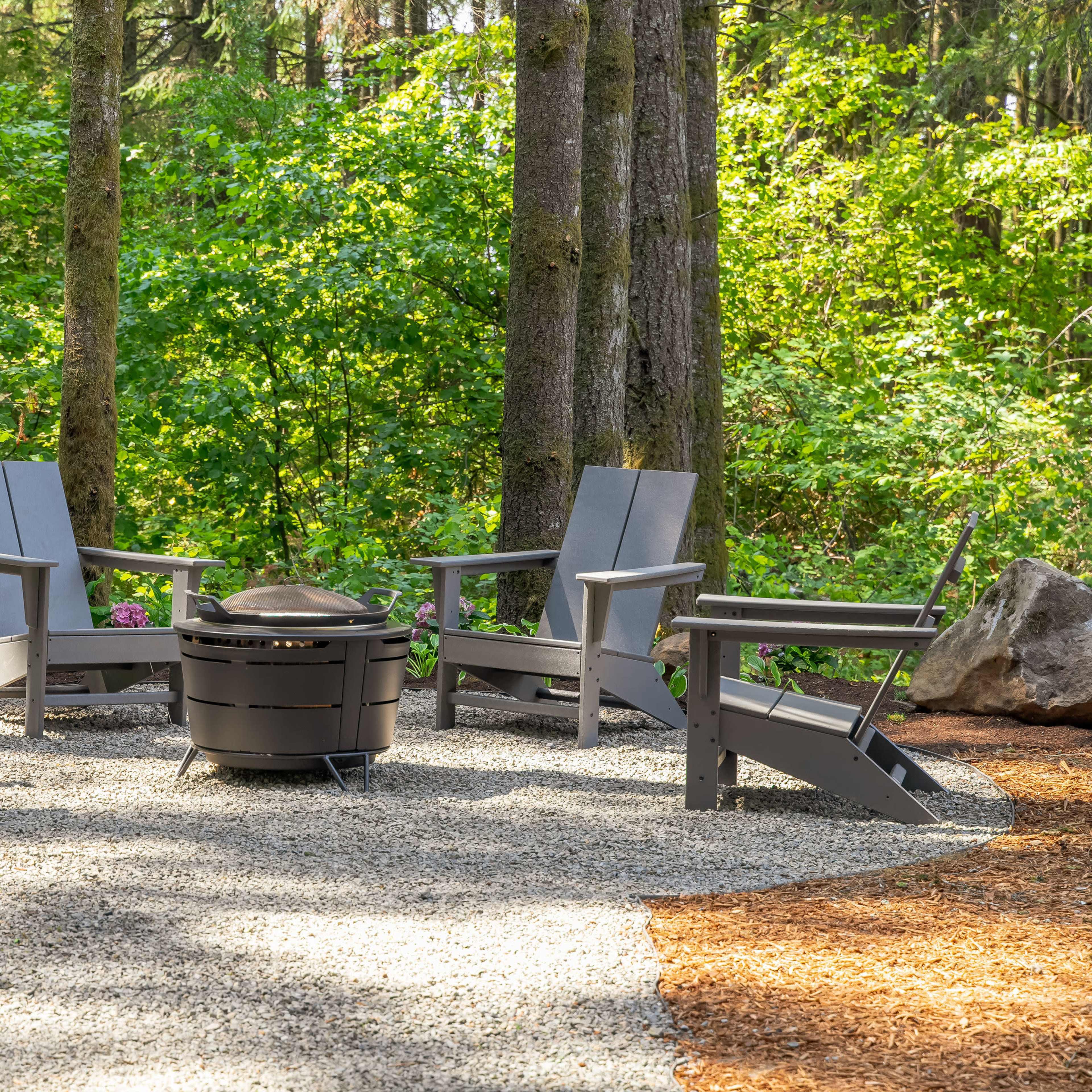 A circular seating arrangement with four gray chairs surrounds a fire pit on a gravel surface, set amidst trees and greenery.