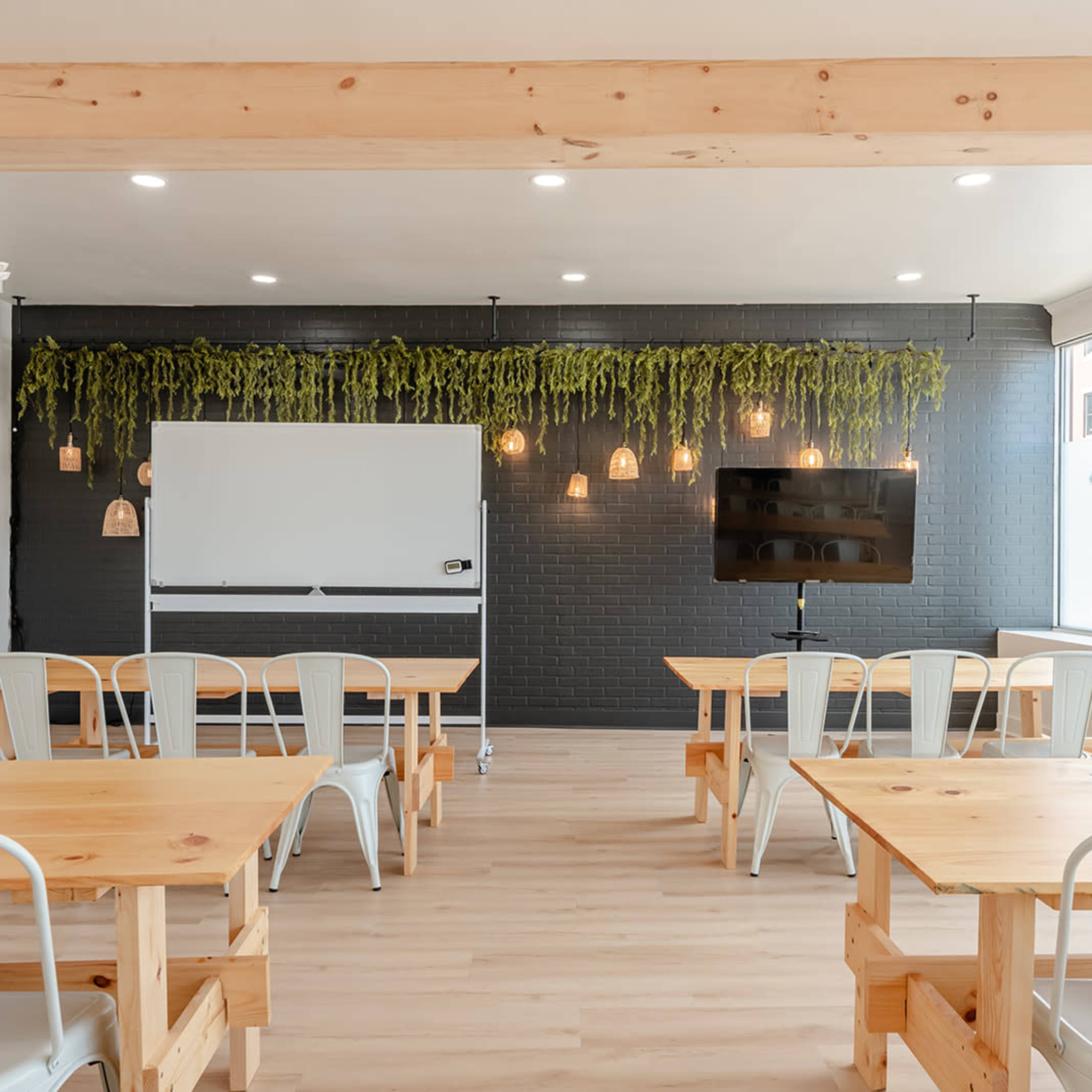 A bright meeting room features wooden tables and chairs, a whiteboard, a television, and greenery hanging from the walls.