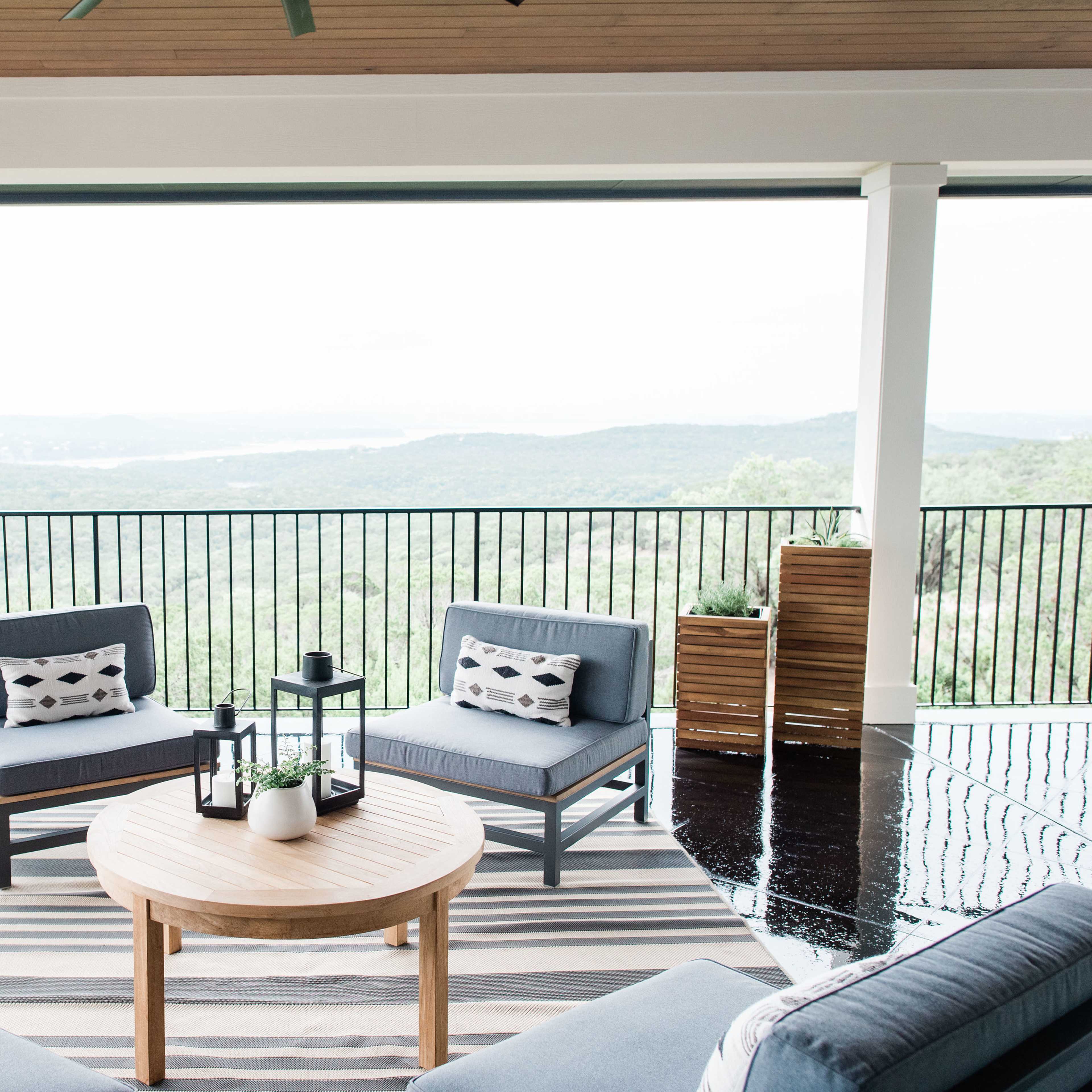 A covered patio with two gray sofas and a round wooden table, overlooking a scenic view of rolling hills and a body of water.