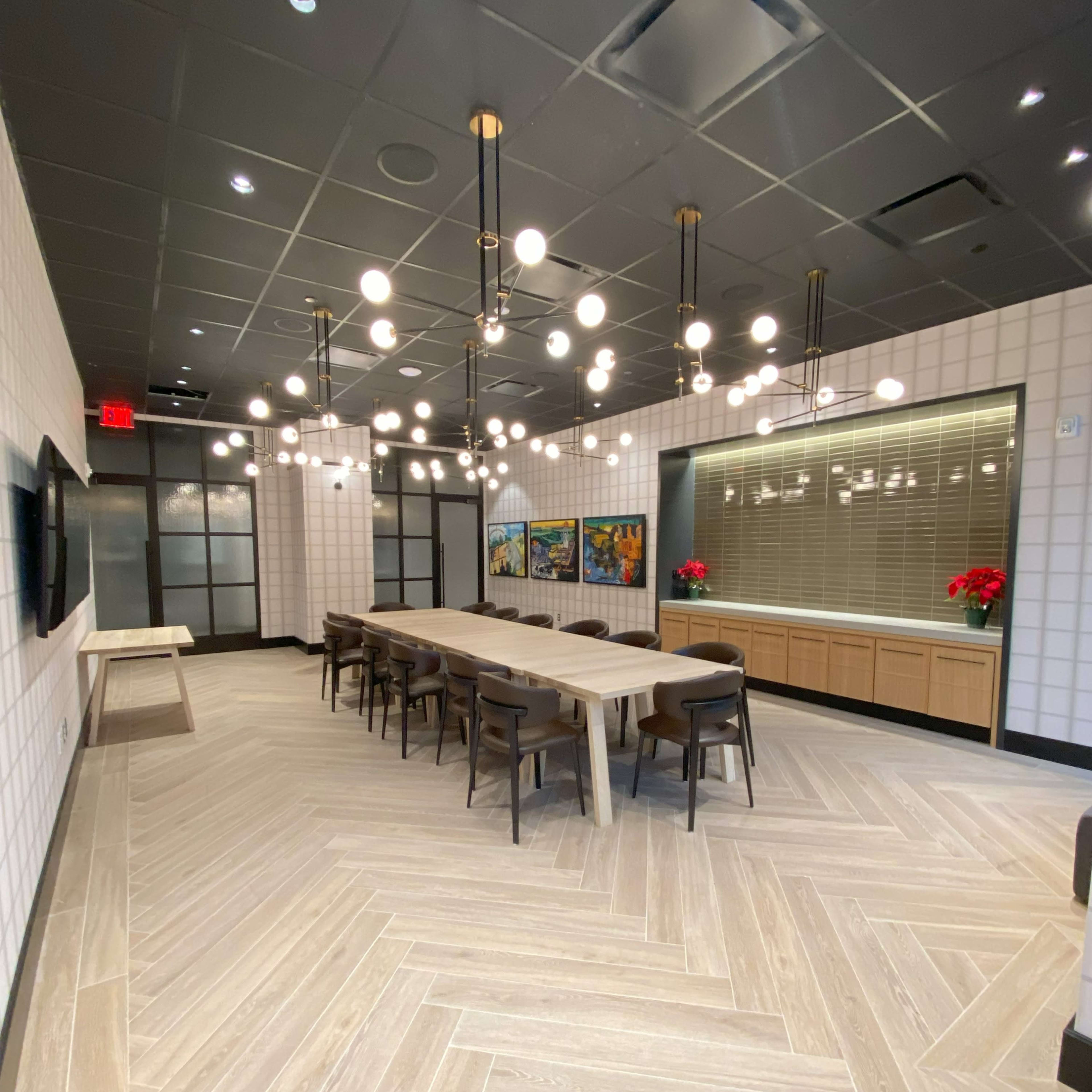 A modern conference room featuring a long wooden table surrounded by chairs, illuminated by spherical pendant lights, with decorative art and potted plants along the walls.
