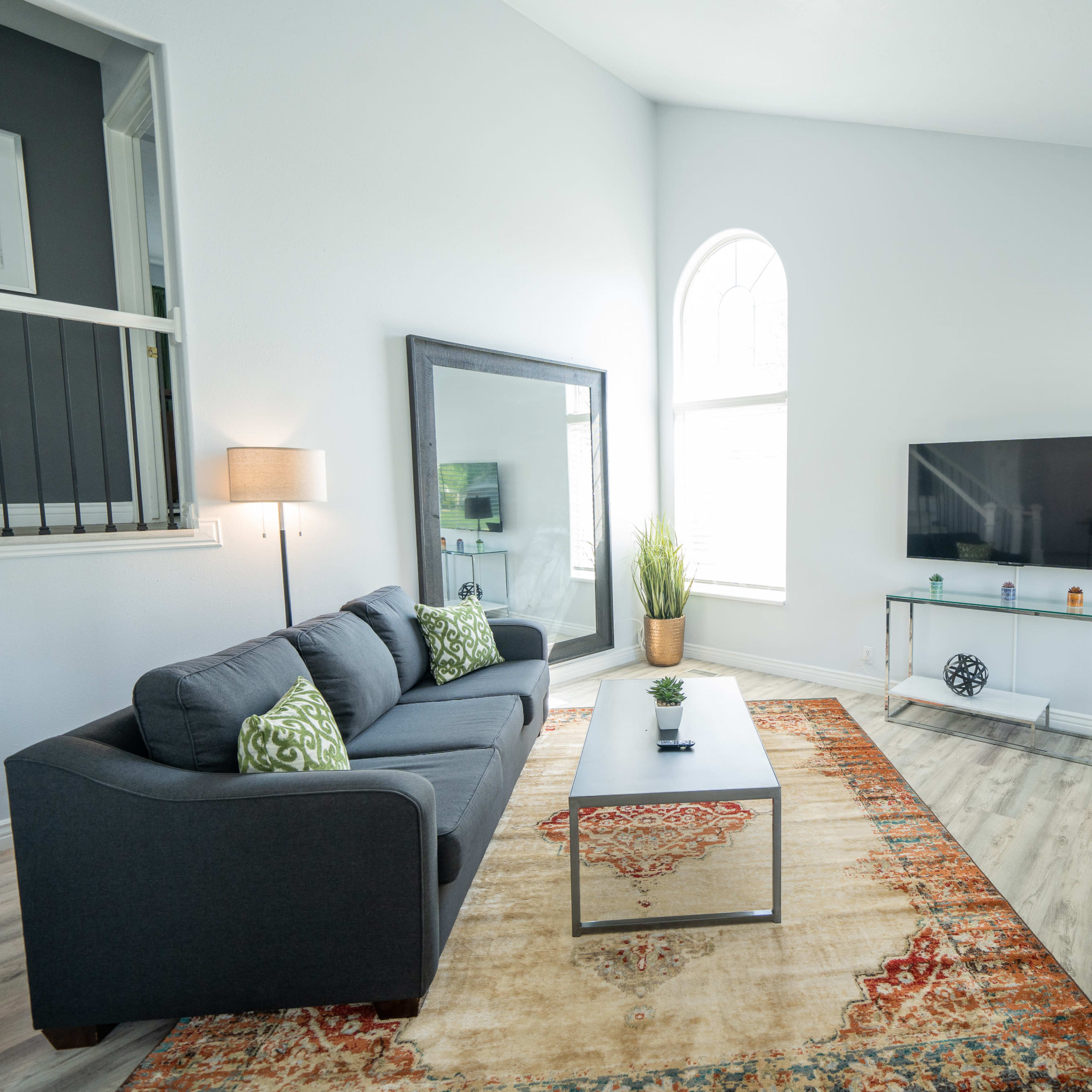 The room features a modern living area with a gray sofa, a green chair, a glass TV stand, and large windows allowing natural light.