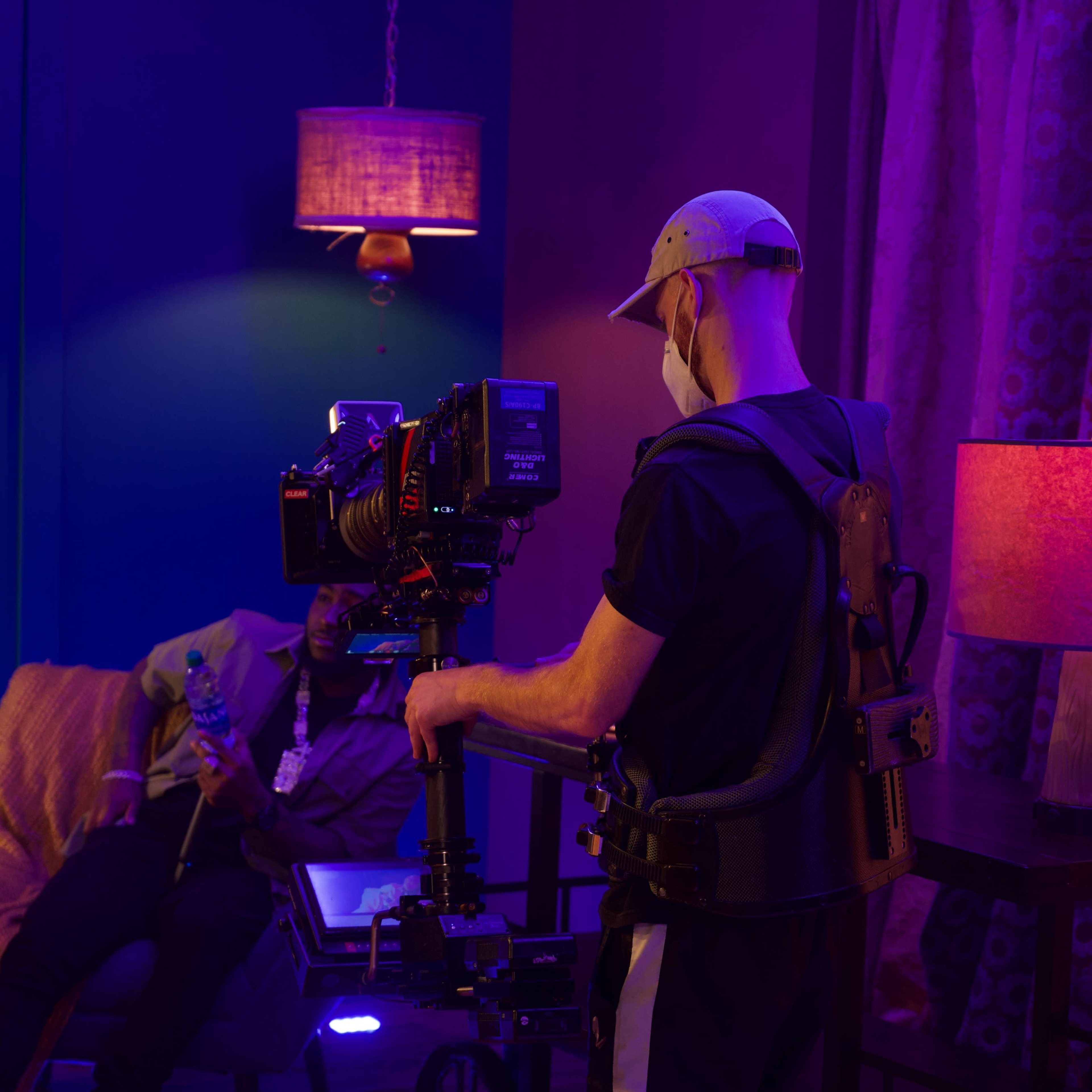 A camera operator uses a rig to film a scene featuring a seated subject in a dimly lit room with colorful lighting.
