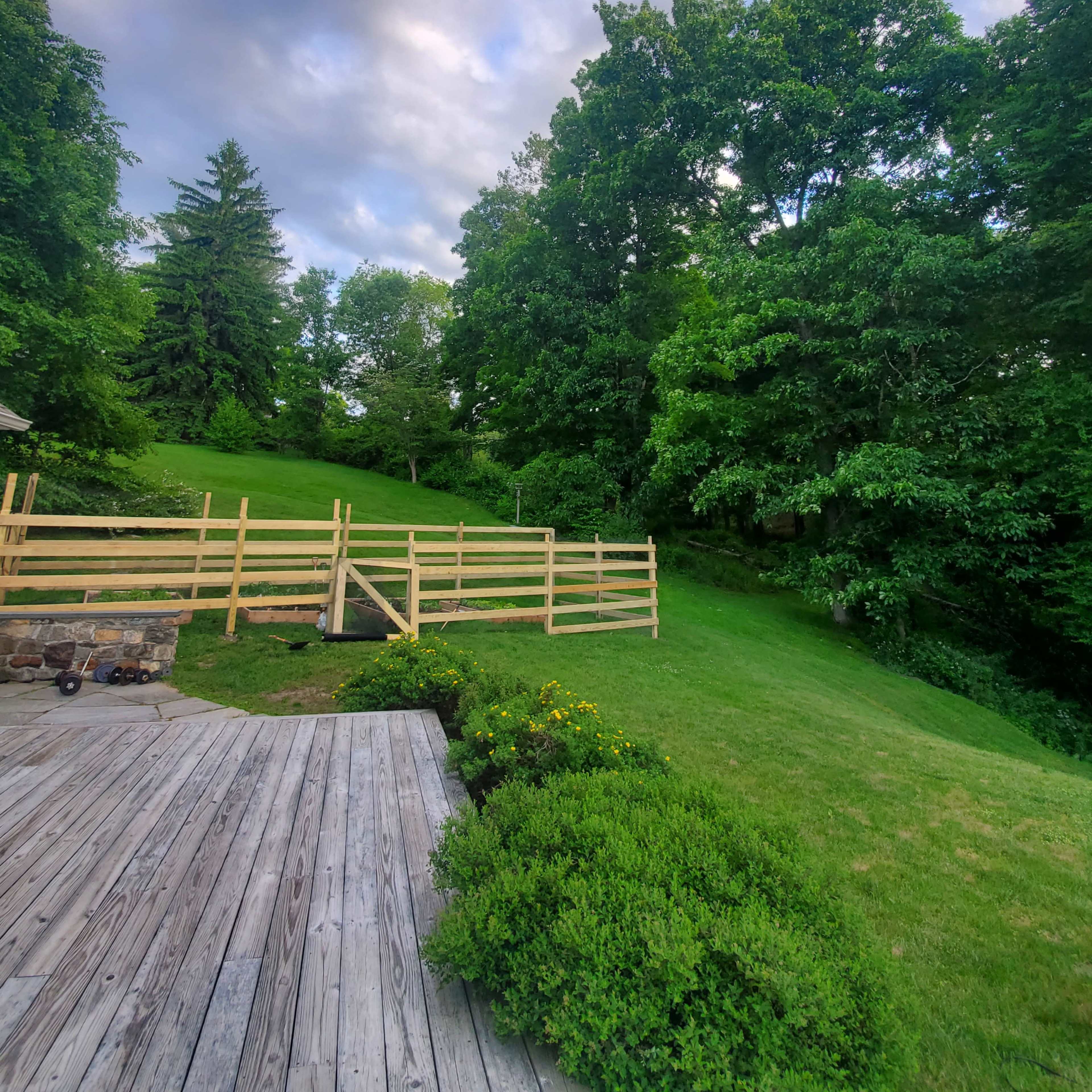 A wooden deck leads to a grassy field bordered by a wooden fence and surrounded by trees under a cloudy sky.