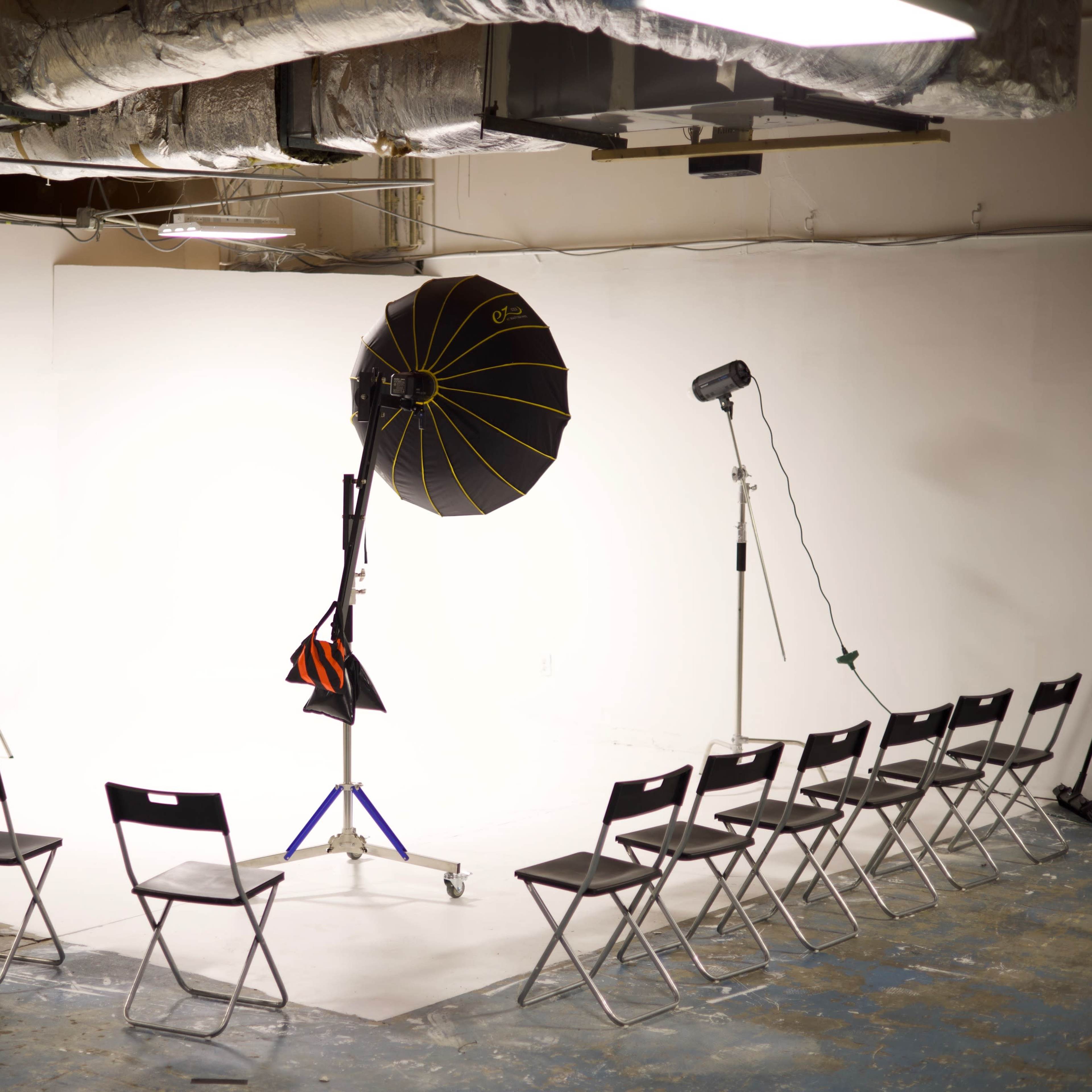 A spacious, well-lit studio is set up with a presentation screen, a large lighting umbrella, and several black chairs arranged in a semicircle.