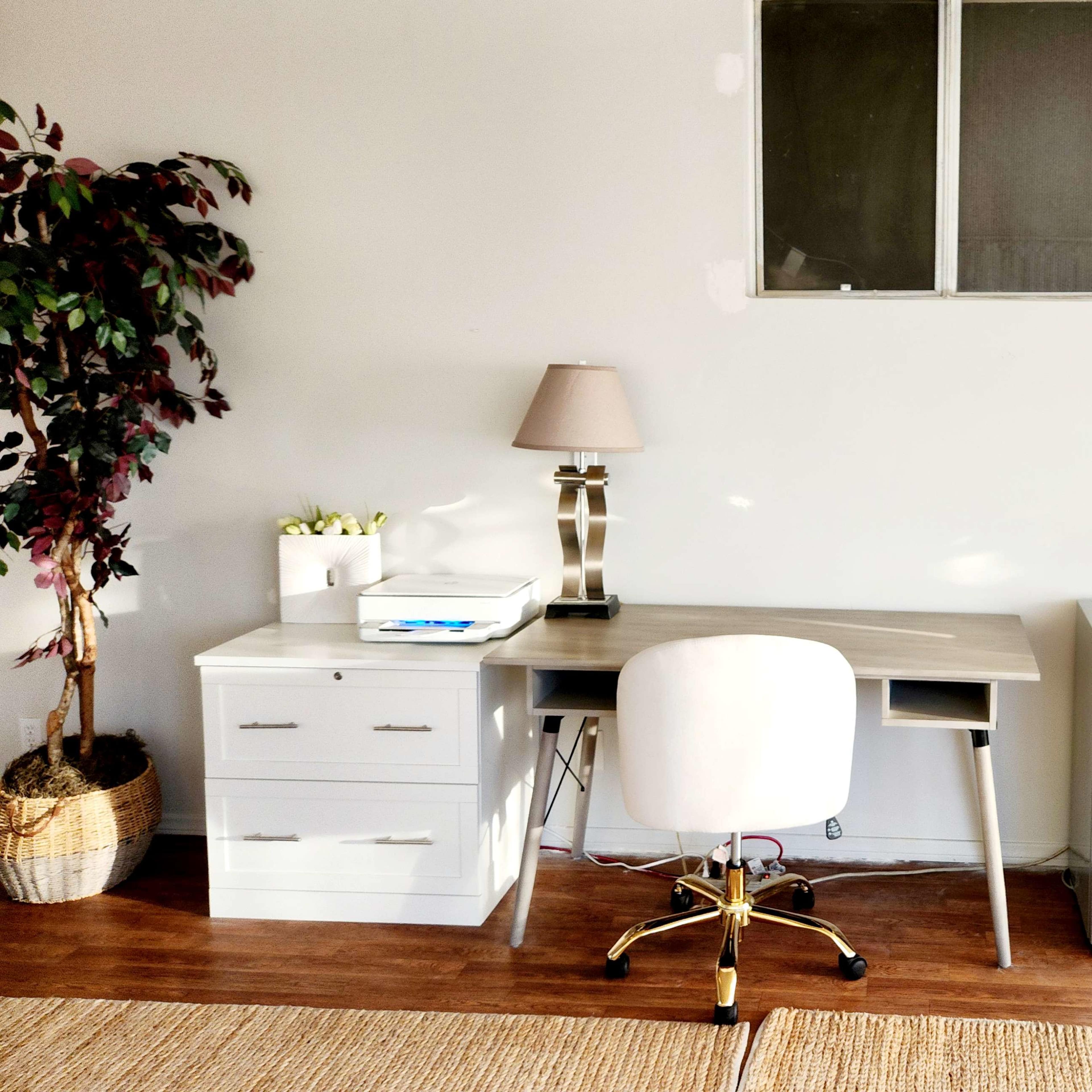 A minimalist home office setup features a wooden desk with a chair, a white filing cabinet, a small refrigerator, and decorative plants in the corners.