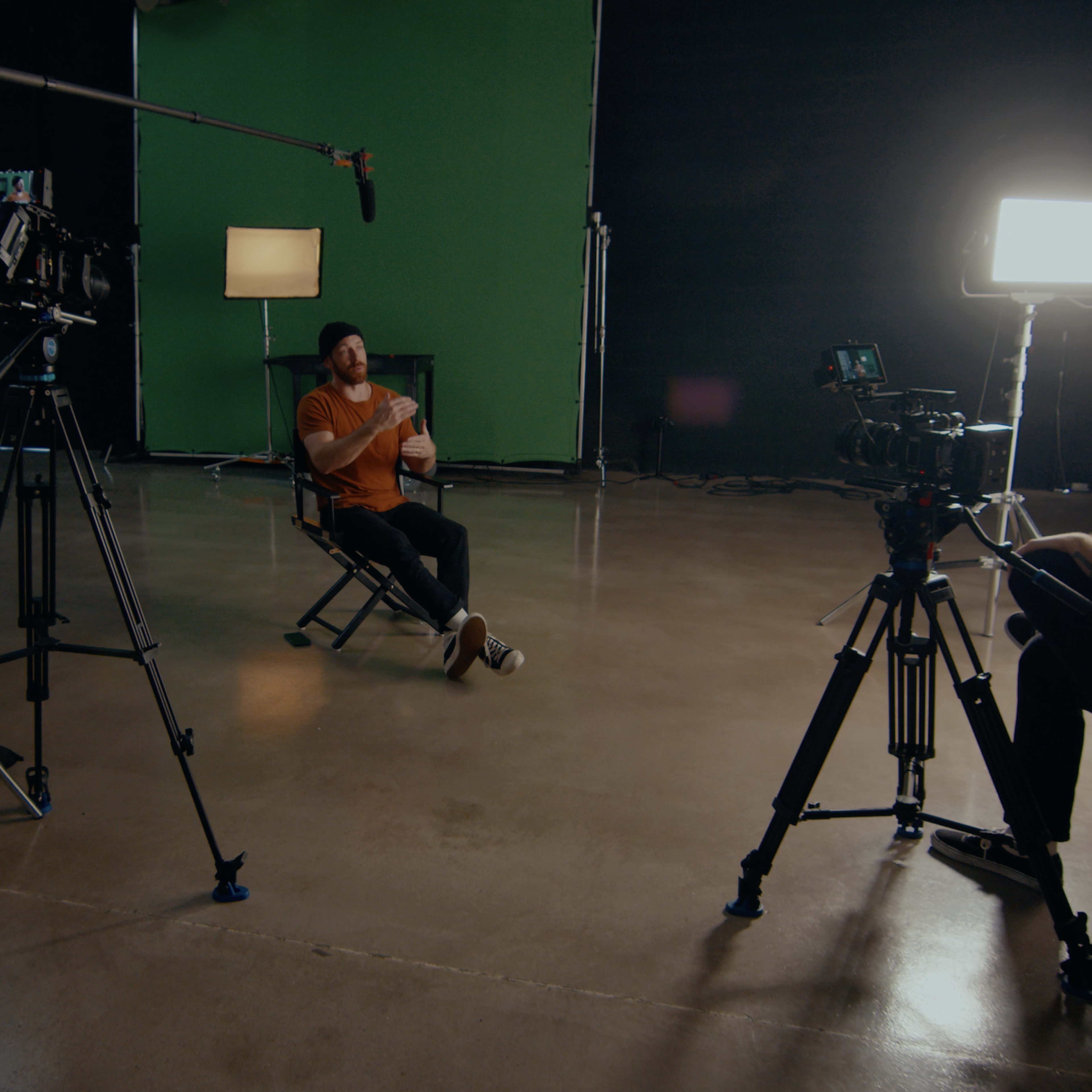 A film set features a seated subject in an orange shirt, surrounded by multiple cameras, a microphone, and a green backdrop.