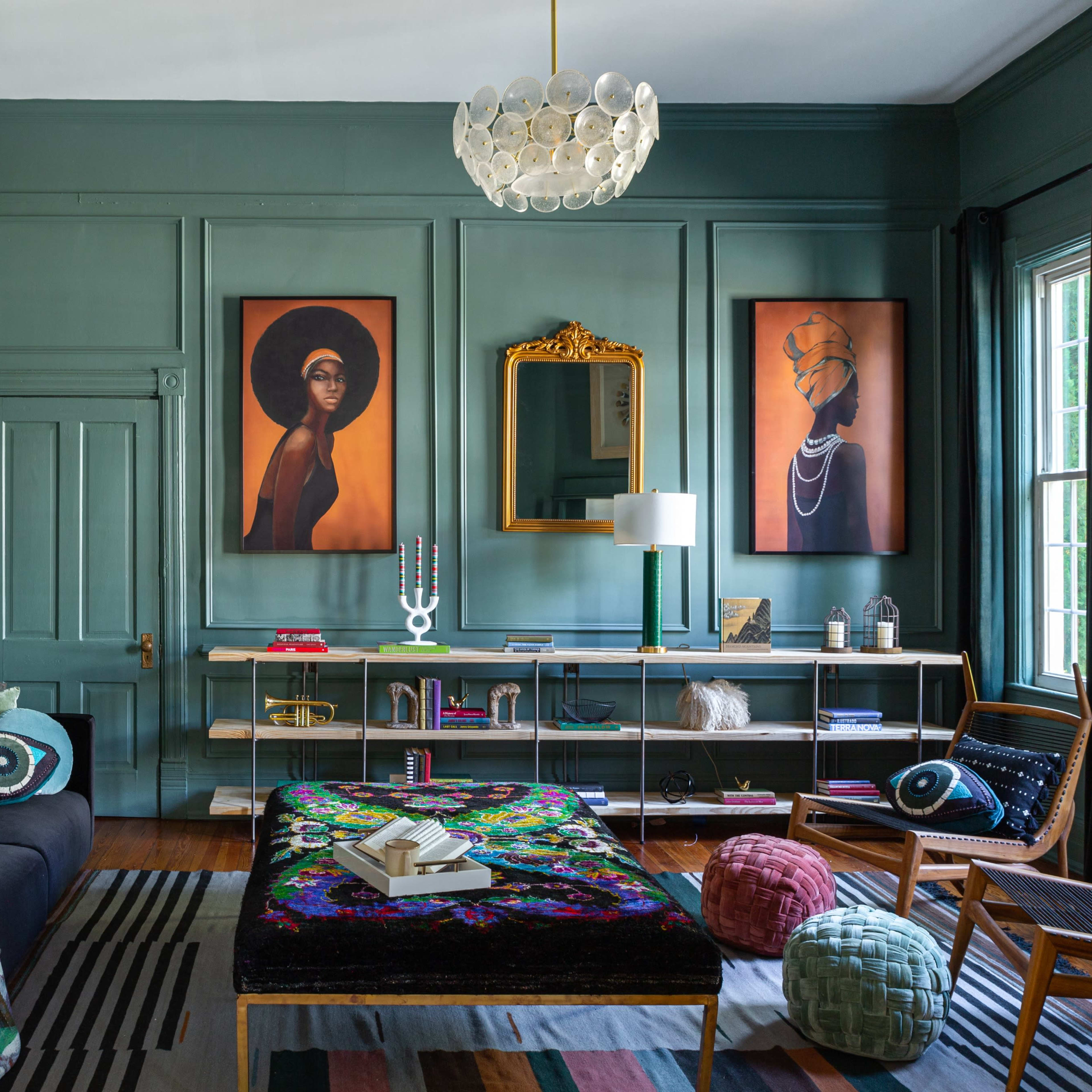 The room features dark green walls, two large artworks, a black sofa with colorful cushions, and a mix of modern and vintage furniture.
