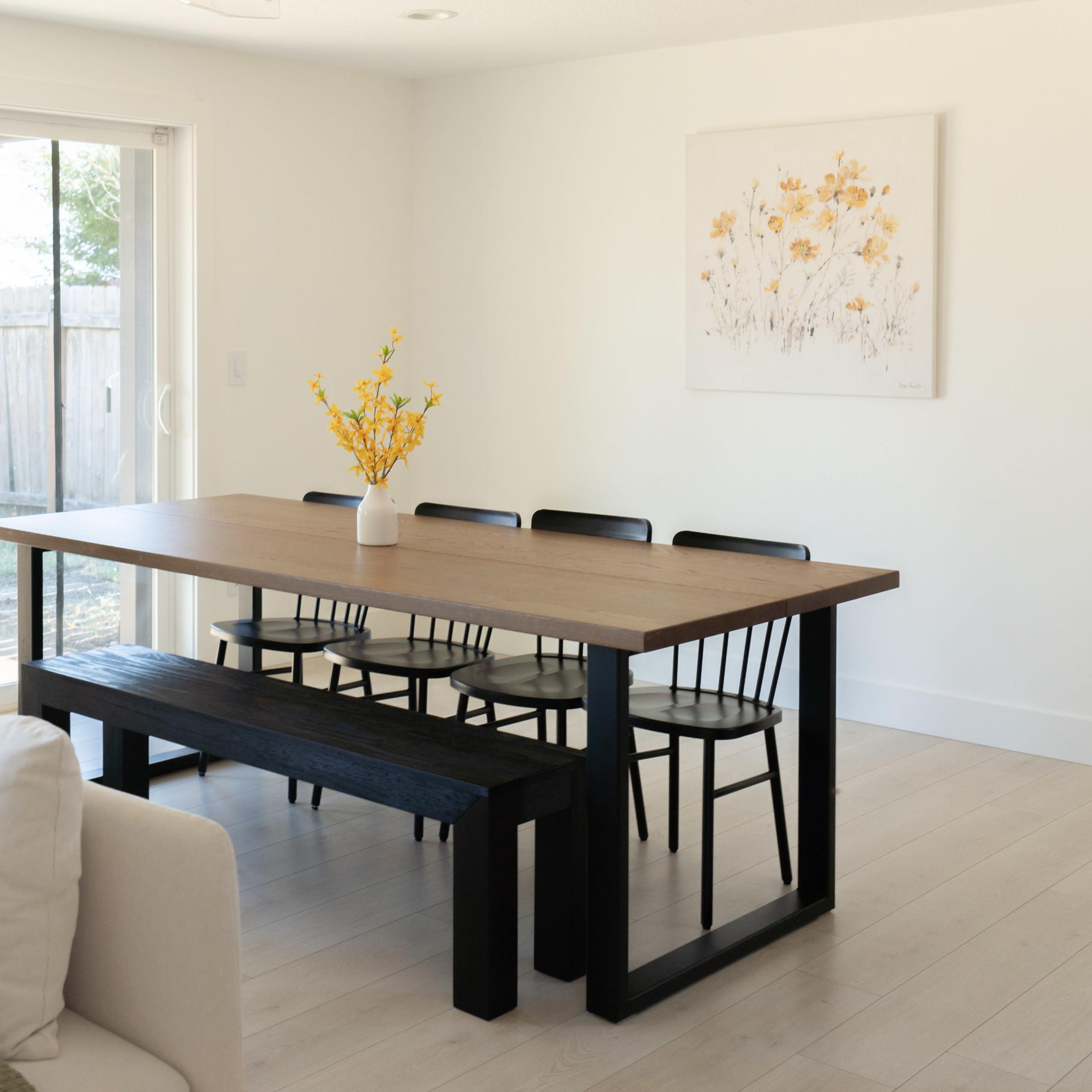 A modern dining area features a wooden table with six black chairs and a bench, complemented by a vase of yellow flowers, against a backdrop of sliding glass doors leading to an outdoor space.