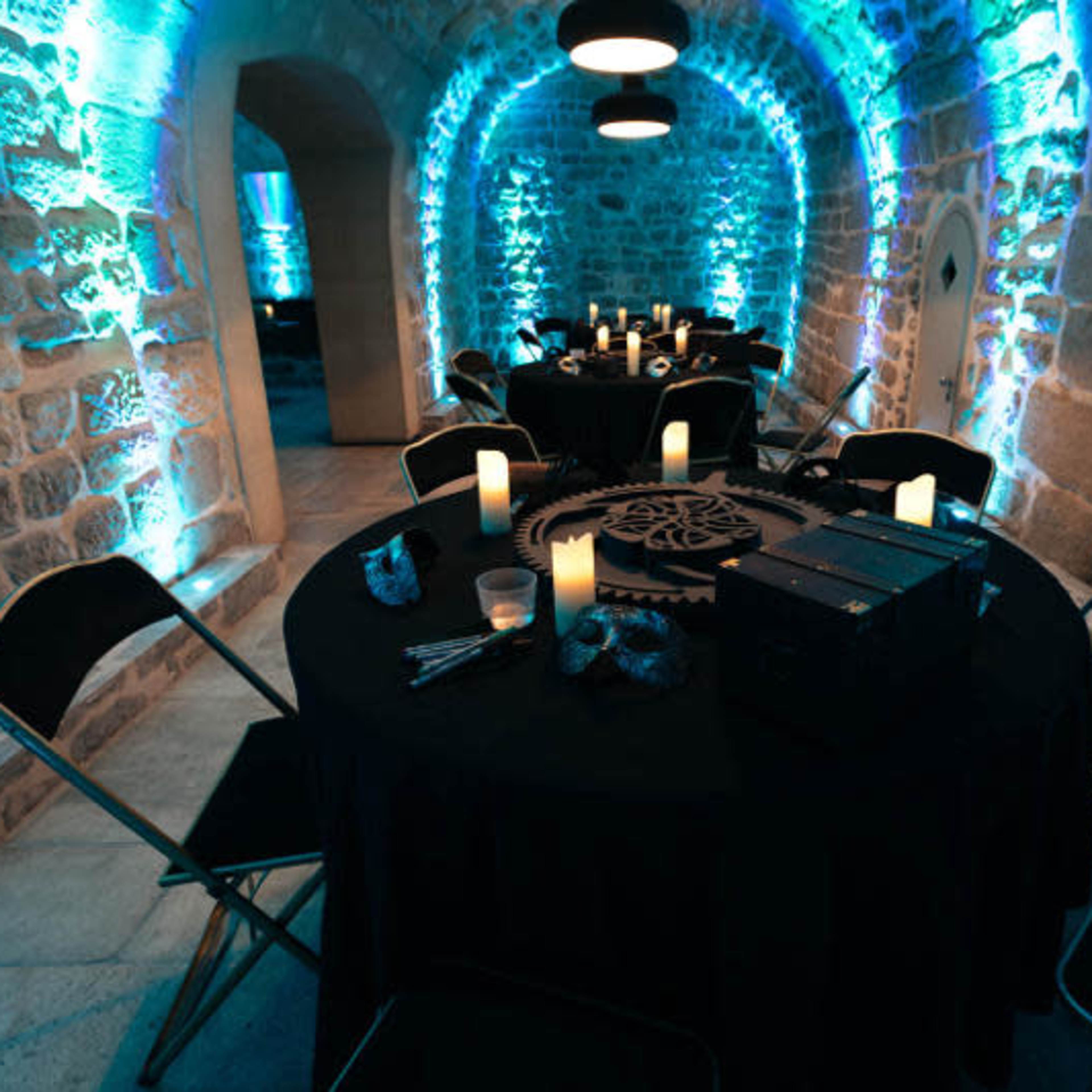 A dimly lit room with stone walls, featuring round tables set with black tablecloths, illuminated by blue lights and surrounded by metal chairs.