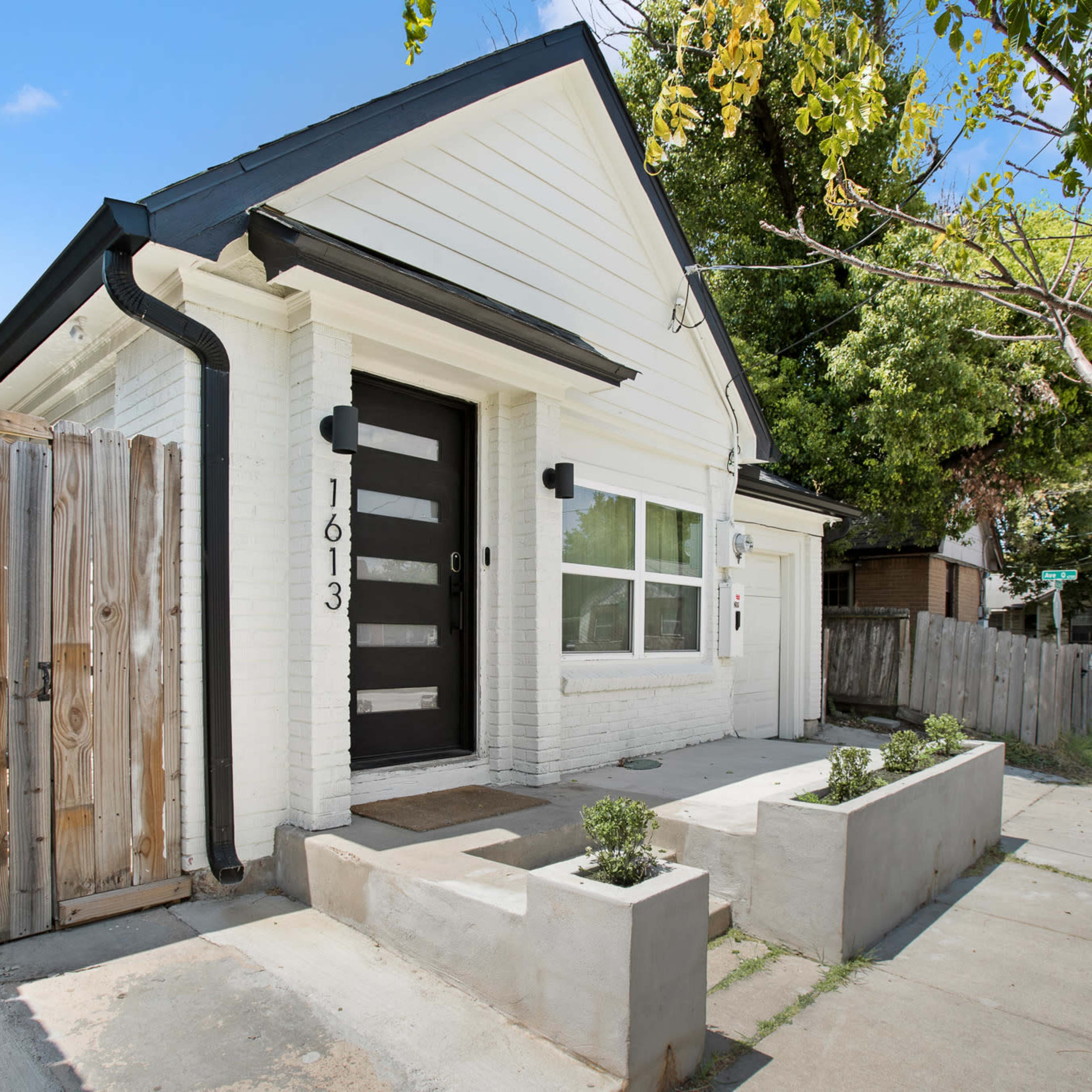 A white, modern-style house with a black door is seen next to a wooden fence and has a small landscaped area in front.