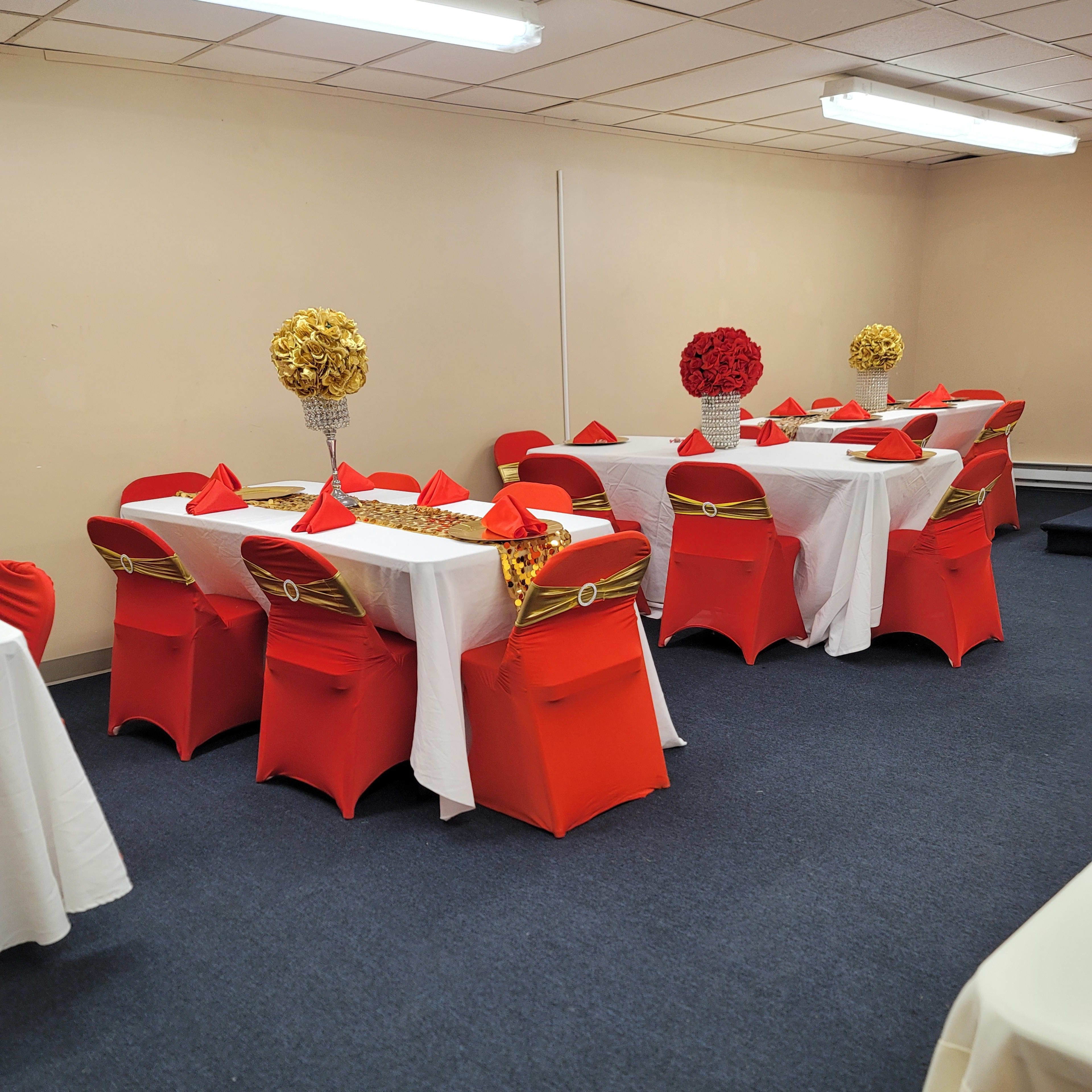 The room features several tables covered with white tablecloths, surrounded by red and gold chairs, arranged for an event celebration.