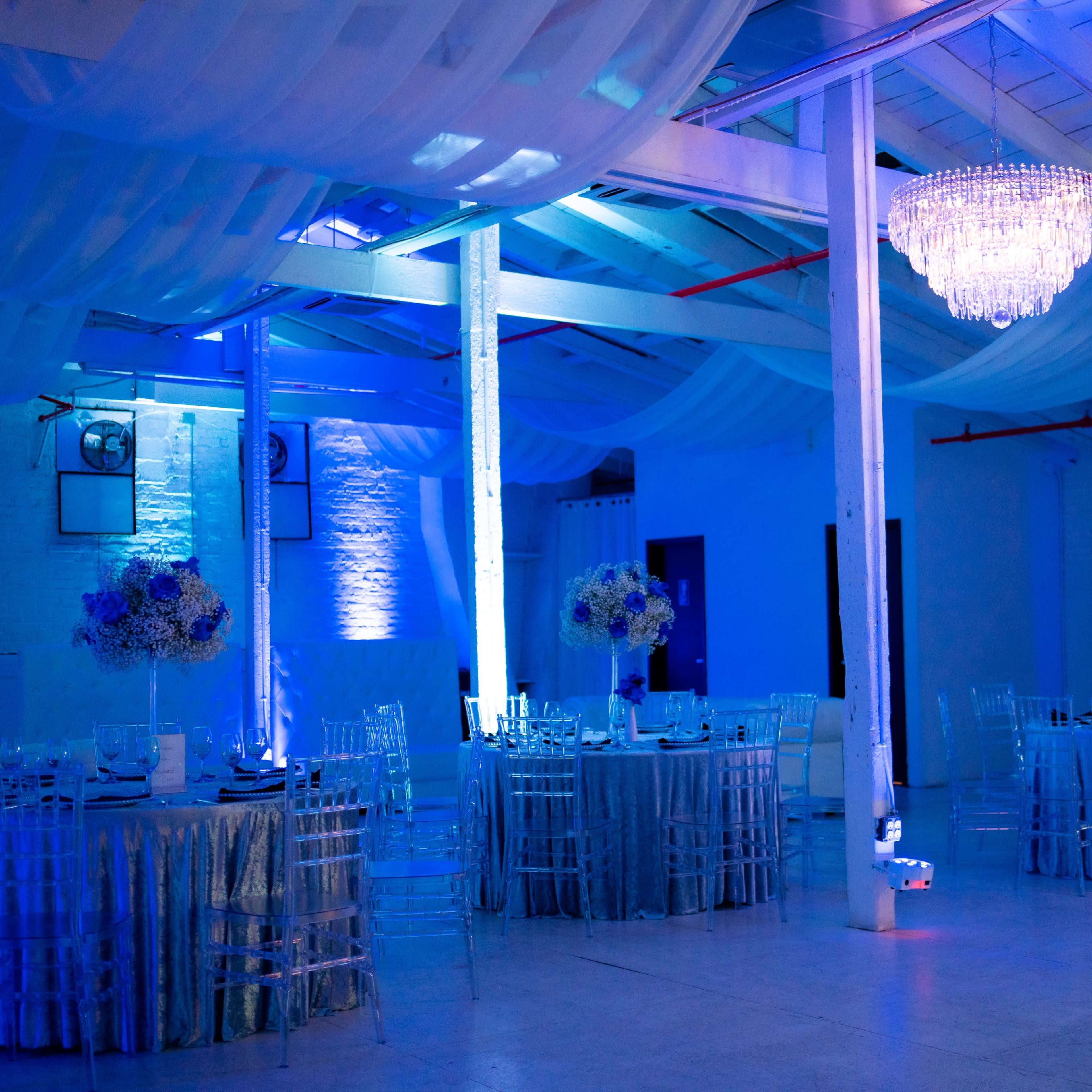 A banquet hall featuring circular tables with silver tablecloths, clear chairs, and a chandelier, all illuminated by blue lighting.