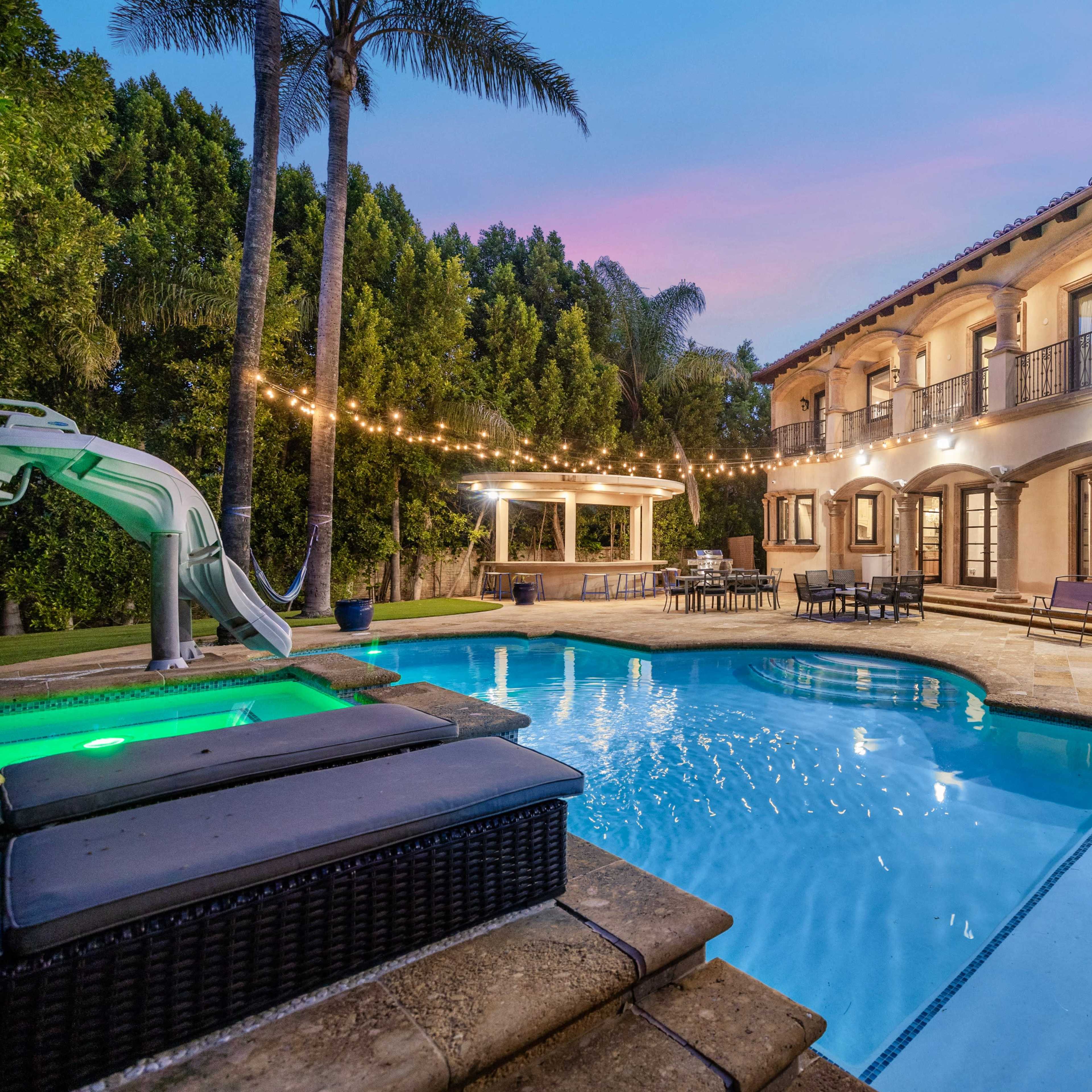 A spacious backyard features a pool with a slide, lounge chairs, and a gazebo surrounded by palm trees and string lights.