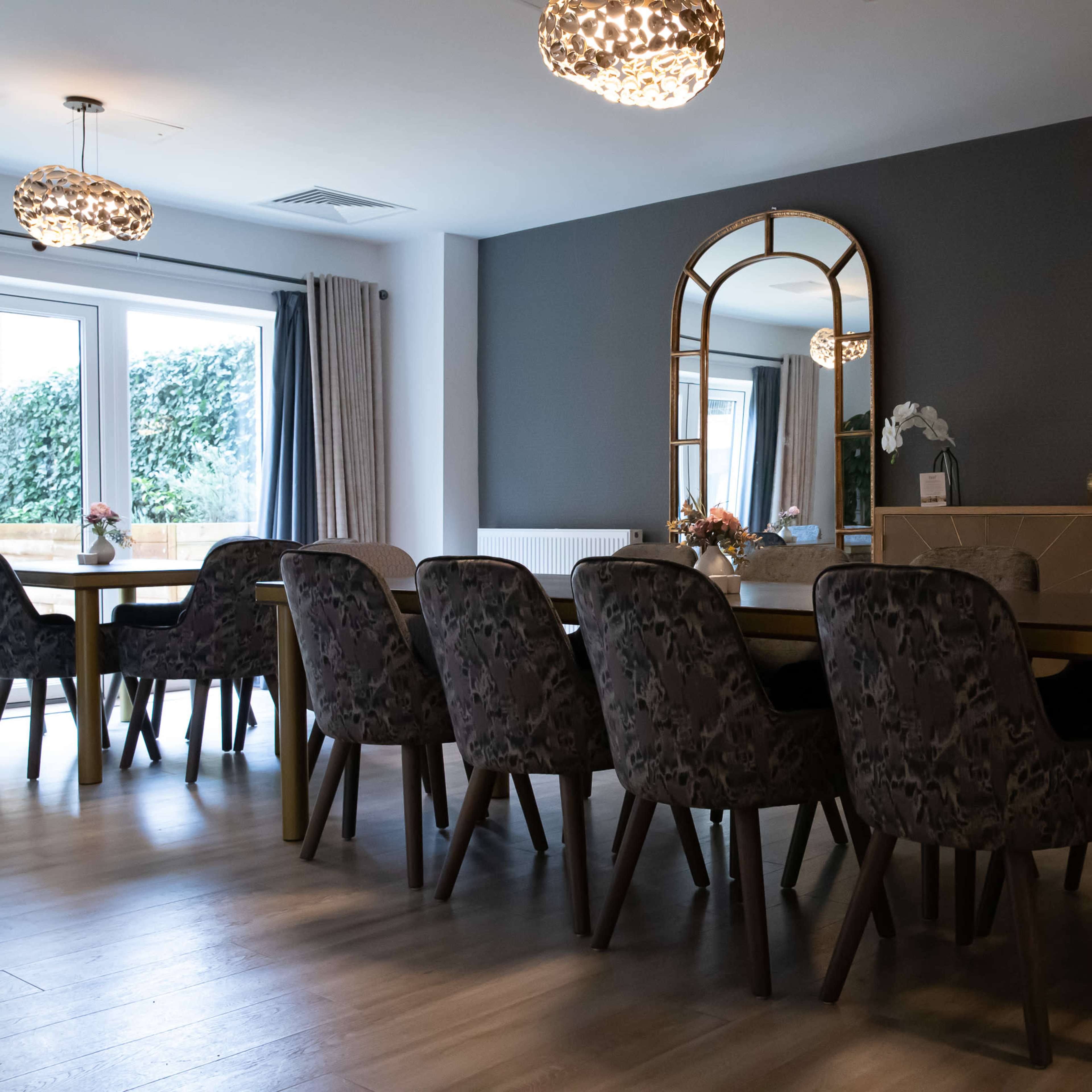A modern dining area features a long table surrounded by upholstered chairs, with large windows and decorative mirrors on the walls.