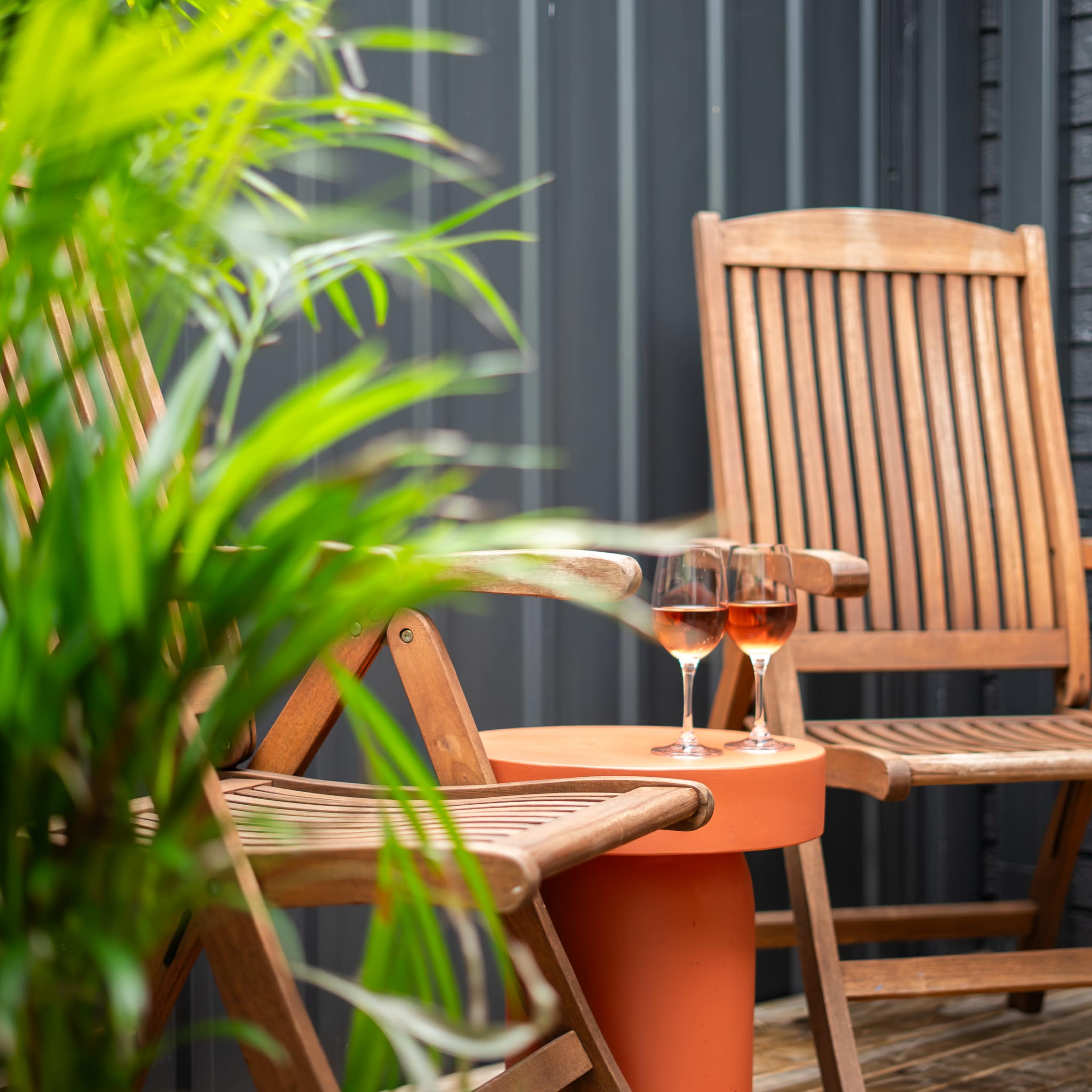 Two wooden chairs are positioned beside a small orange table with two glasses of rosé wine, set against a backdrop of green foliage.