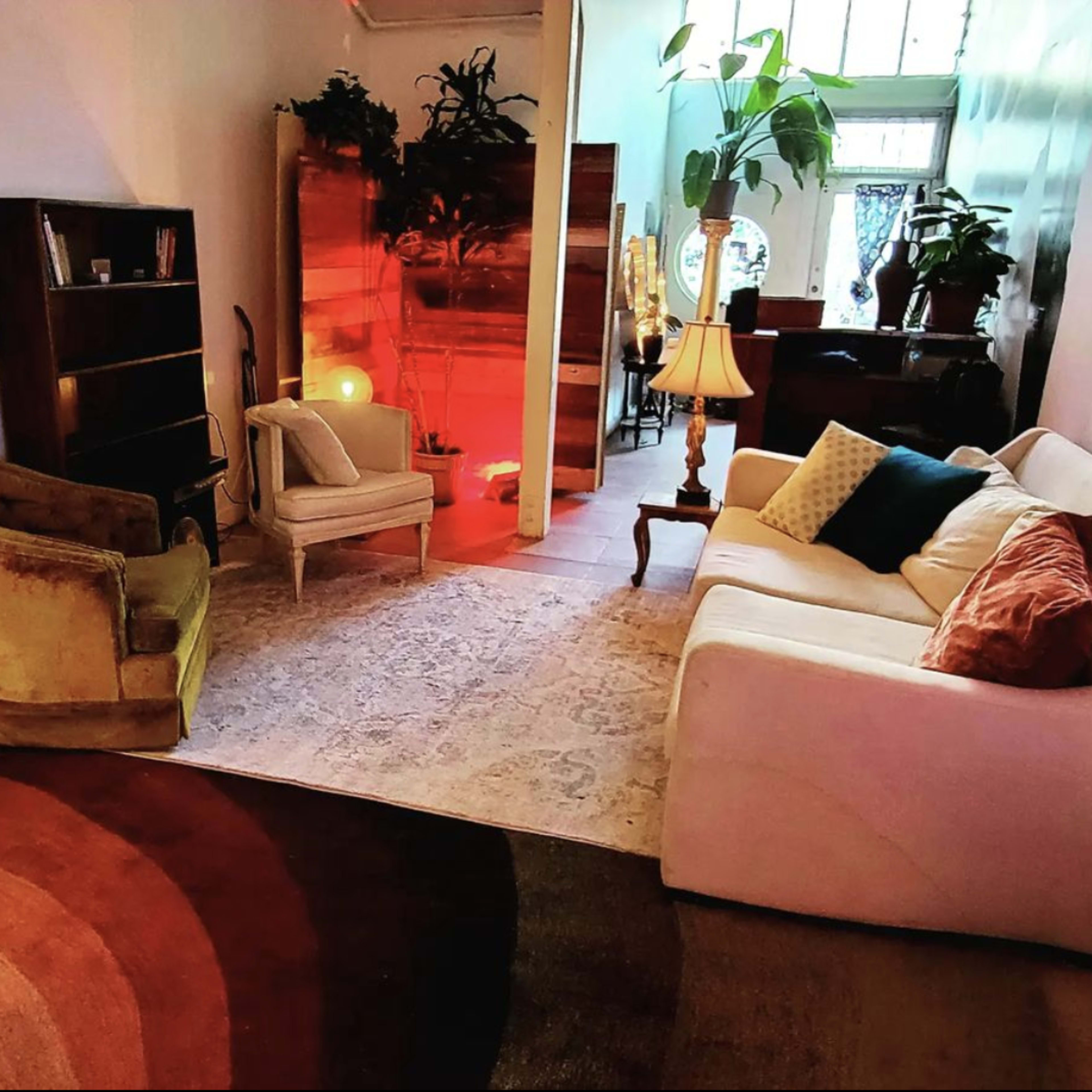 A living room with two armchairs and a sofa arranged around a circular rug, with plants and a bookshelf in the background.