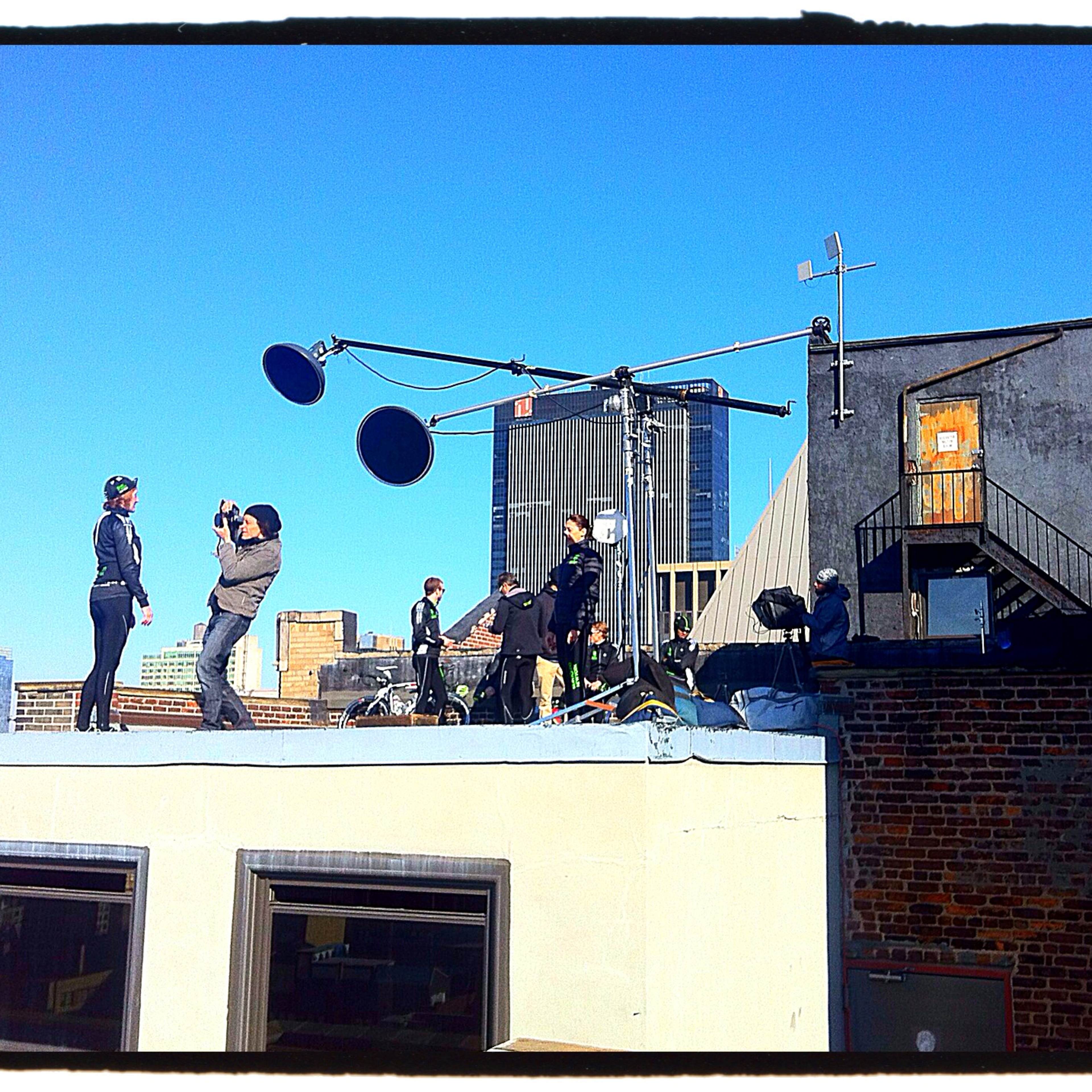 A film crew is shooting a scene on a rooftop with city buildings in the background and equipment set up nearby.