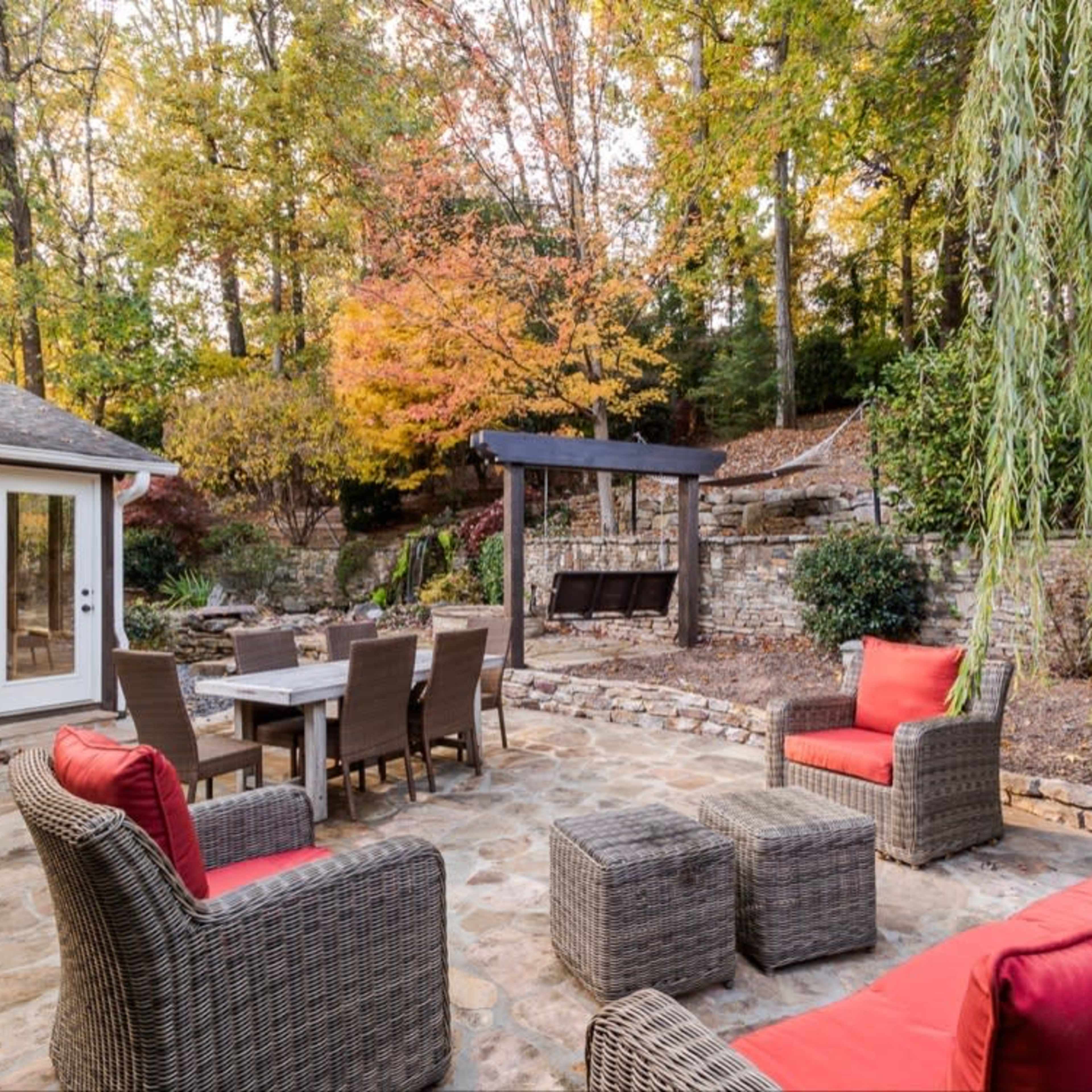 A stone patio with several seating options, including red-cushioned chairs, a dining table, and a pergola, surrounded by trees displaying autumn foliage.